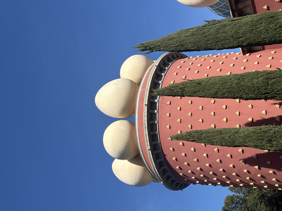 The image showcases a portion of the Salvador Dalí Theatre-Museum in Figueres, Spain. A cylindrical, reddish structure with repetitive, small, golden ornaments is prominent. Rows of elongated, green trees encircle the cylinder. Large, egg-shaped sculptures adorn the side facing the clear blue sky. Architectural details of the museum are visible on top.