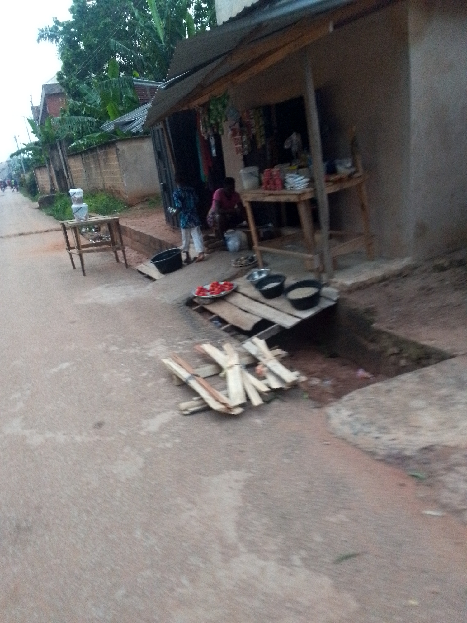 The image captures a roadside scene in what appears to be a rural or semi-urban setting, consistent with Orlu, Nigeria. A small, open-front shop or stall, constructed with wooden posts and a corrugated metal roof, stands on the right side of a dusty, unpaved road.
Two individuals are visible near the shop. One person is sitting inside or just outside the stall, wearing a reddish-pink top. Another person is standing nearby, dressed in a patterned blue and white top and white trousers.
The shop displays various items, with packaged goods visible hanging and on a wooden table. On a wooden platform bridging a drainage ditch in front of the shop, there's a plate filled with sliced red items, possibly tomatoes or peppers, alongside two black buckets containing what appears to be grains or produce. Bundles of light-colored wooden planks or sticks are stacked on the ground next to the ditch. Further to the left on the roadside, a smaller wooden table holds clear stacked containers.
The road itself is a light, dusty color with some wear. Lush green trees and banana plants line the background and a low concrete wall to the left, indicating a tropical environment. The lighting is soft and diffused, suggesting either early morning, late afternoon, or an overcast day. No specific text or branding is clearly legible anywhere in the image. The scene depicts a typical local market setup or community store.