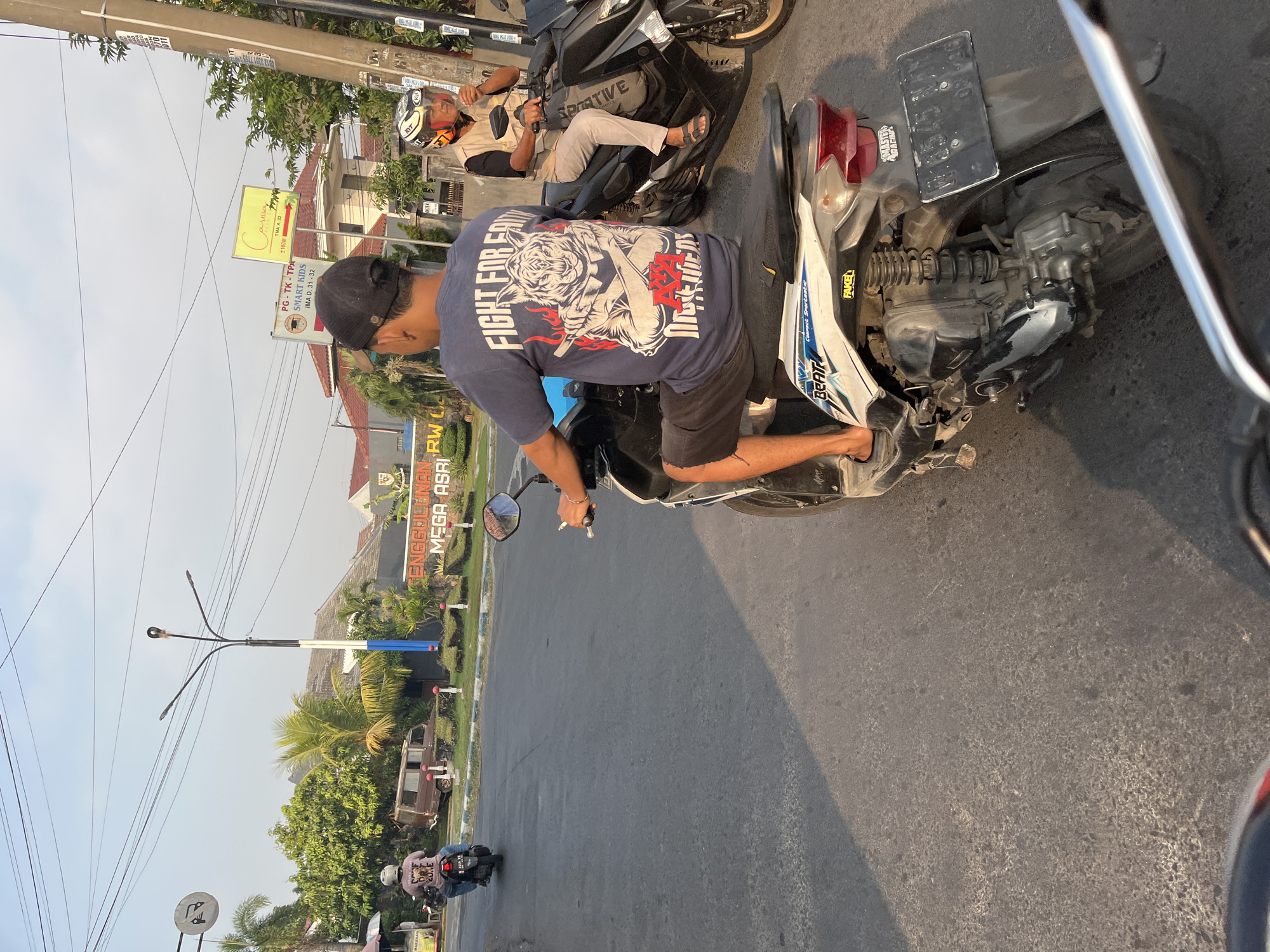This is a medium shot of a street scene in Tenggulunan, Indonesia, taken on a sunny day. The perspective is from behind the viewer's own motorcycle, looking forward at other motorcycles and traffic on a paved road. In the foreground, the handlebars and seat of the viewer's motorcycle are visible.
Dominating the mid-ground is a man wearing a dark blue t-shirt with a large graphic of a tiger with crossed arms and flames, and black shorts. He is riding a white and blue scooter. To his right, another man wearing a light-colored vest over a dark shirt and a white and orange helmet is riding a black motorcycle. He is holding a cigarette in his right hand.
In the background, several buildings are visible, including what appears to be a school with signs that read "PG-TK-TPA SMART KIDS" and "TMA A. 22". Another sign says "ENGGULUNAN MEGA ASRI RW 6". A brown van is parked to the left of the road. Palm trees and other greenery are scattered throughout the scene. Overhead, numerous electrical wires crisscross the sky. The overall impression is a typical urban street with ongoing traffic.