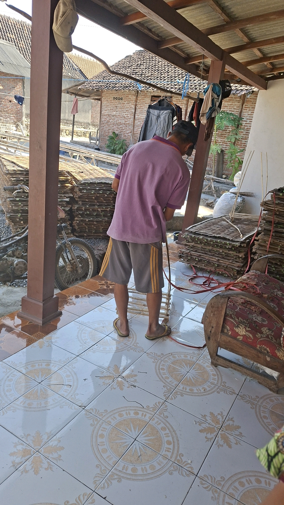 In the city of Sambeng, Indonesia, a man stands on a tiled patio under a rustic wooden-roofed structure. He is wearing a purple t-shirt and gray shorts with yellow stripes. He appears to be working with a red hose or rope that snakes across the floor. To his left, a stack of what appears to be woven mats or thin wooden planks is piled high. A motorcycle is parked nearby, its front wheel visible. The background reveals brick buildings with tiled roofs, suggesting a residential or workshop area. Several items of clothing are hanging on a line under the roof. To the man's right, another stack of similar woven materials is visible, along with a large wooden object with patterned upholstery, possibly a bench or decorative item. The tiled floor features a circular, star-like pattern in a light yellow hue.