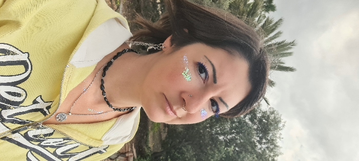 A medium shot depicts a single individual, appearing female, outdoors in the Mate Asher Regional Council, Israel. The person has brown hair and is looking slightly to the camera's right with a neutral expression. Both cheeks feature glitter makeup. The individual wears blue eyeliner/mascara, a silver stud on the right nostril, and a silver septum ring. The left ear displays multiple earrings, including a dangling one with a crescent moon charm and chains. They are dressed in a yellow hooded sweatshirt with a partial zipper and white trim, featuring a dark graphic print on the chest. Necklaces include a black beaded choker and a longer silver chain with a circular "tree of life" pendant. Glitter is also visible on the chest below the neckline. The background consists of an overcast sky with grey clouds, green foliage including palm fronds, and olive trees. A light-colored structure is partially visible in the lower right background.