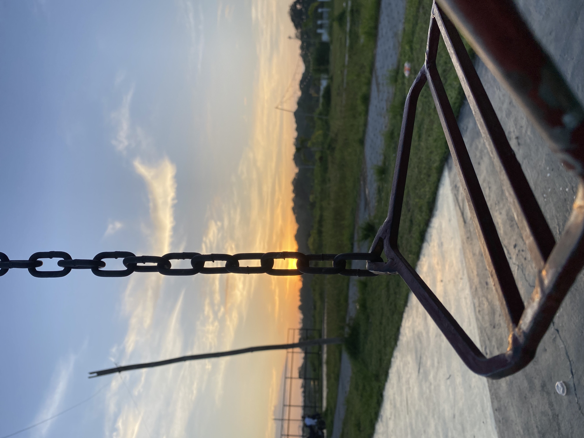 The image depicts a close-up of a rusty swing set chain hanging in the foreground, with a sunset visible in the background. The swing seat itself is partially visible, made of metal bars and worn paint. Beyond the swing set, there is a grassy field with a dirt path and some fencing in the distance. Several trees form a dark silhouette against the horizon. The sky is a mix of blue and orange hues, with wispy clouds illuminated by the setting sun. The overall setting appears to be a public park or an outdoor recreational area, possibly located in Davao City, Philippines, given the vegetation and lighting conditions consistent with a tropical sunset. There are no visible people in clear focus.