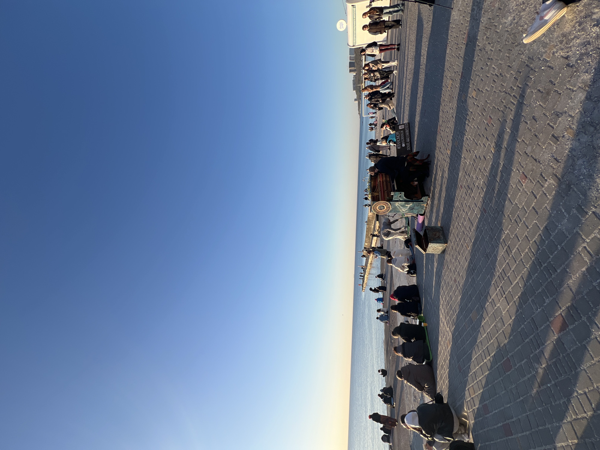 The image captures a serene outdoor scene on a paved promenade next to the sea in Odesa, Ukraine, under a clear blue sky. The sky transitions from a deep blue overhead to a lighter shade near the horizon, meeting the calm, reflective waters of the sea.
A large group of people is present, predominantly seated or kneeling on the patterned grey pavers, facing towards the water. Their postures suggest a shared activity of prayer, meditation, or quiet contemplation. Long shadows stretch across the promenade, indicating either early morning or late afternoon sun. Some individuals are standing further back from the water's edge.
In the foreground, a person is seated near what appears to be a barrel organ or a similar mechanical instrument. Adjacent to this, a black sign with white text is clearly visible, displaying: "#ПЯНОМОРЕ", "INST: @DANISHOVKA", and a donation number: "DONATE: +44 7444 095870". Further along the promenade, another sign, partially visible, advertises "НАТУРАЛЬНОГО МОРОЗИВО" (Natural Ice Cream). The overall impression is one of peaceful communal gathering by the sea.