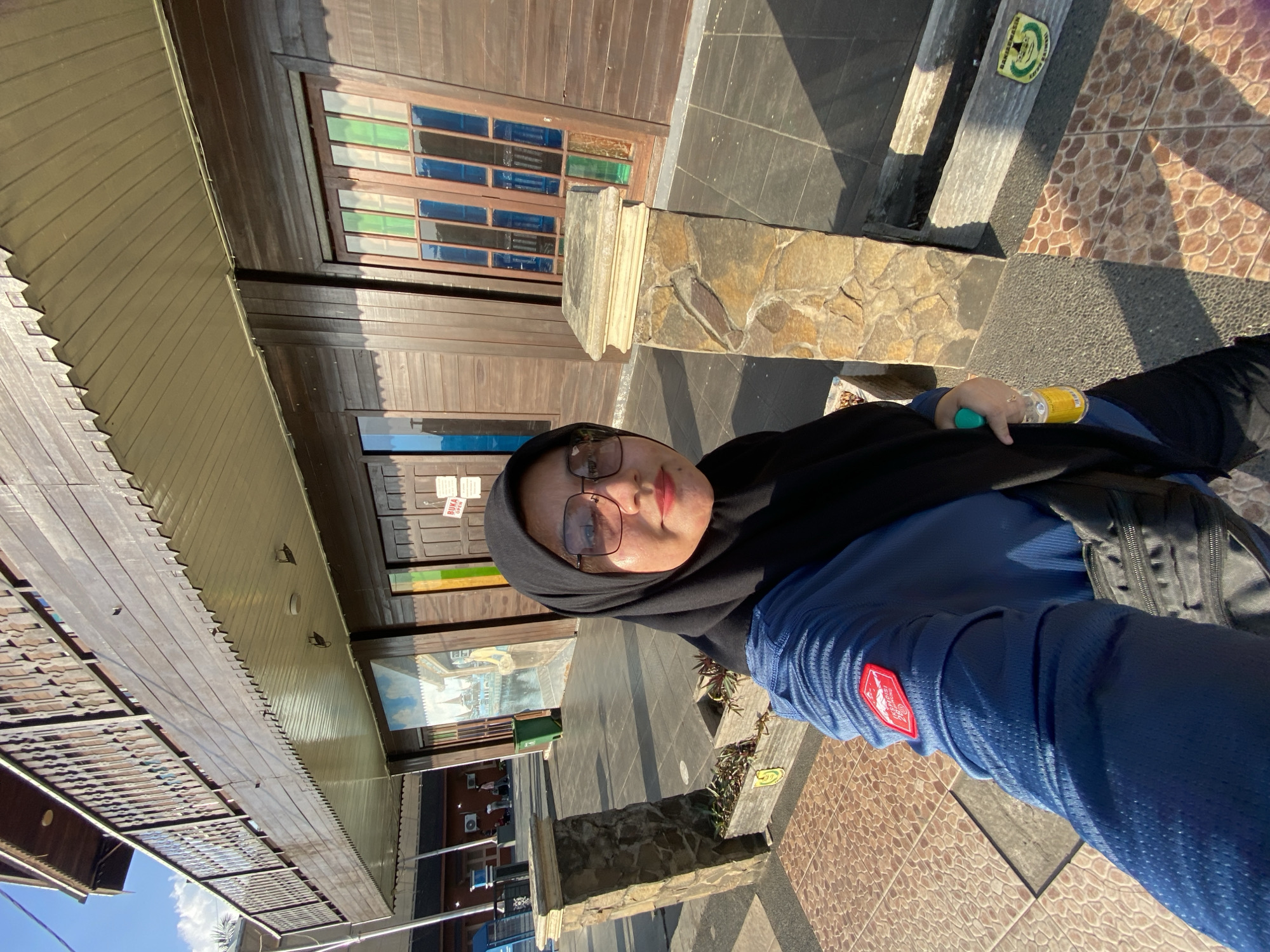 A woman wearing a black hijab and glasses, a blue long-sleeved shirt, and dark pants is taking a selfie outdoors. She is standing in front of a building with brown wooden siding and balconies. The building has several windows, some of which are stained glass. There is a mural on the side of the building to the left of the woman, and a green trash can is visible to the left of the mural. The ground is paved with tiles. The woman is holding a yellow bottle in her right hand. A black bag is slung over her left shoulder. A red badge with white lettering is visible on the sleeve of her shirt. Two stone pillars are present in the foreground. The environment appears to be sunny, with shadows cast on the ground. The scene is set in the city of Banjarmasin, Indonesia, suggested by the presence of the "BANJAR MASA KINI" logo on a plaque on a planter box to the right of the frame.