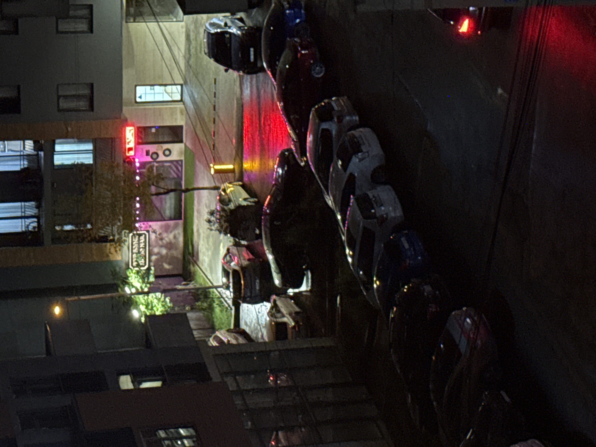 A nighttime, high-angle exterior shot shows a dark, wet street in front of a modern building in Tbilisi, Georgia. Cars are parked on the street, some with their headlights on, casting reflections on the wet pavement. A neon sign glows in front of a doorway on the building. The building appears to be a multi-story apartment complex or commercial building. The sky is dark, indicating it is nighttime.