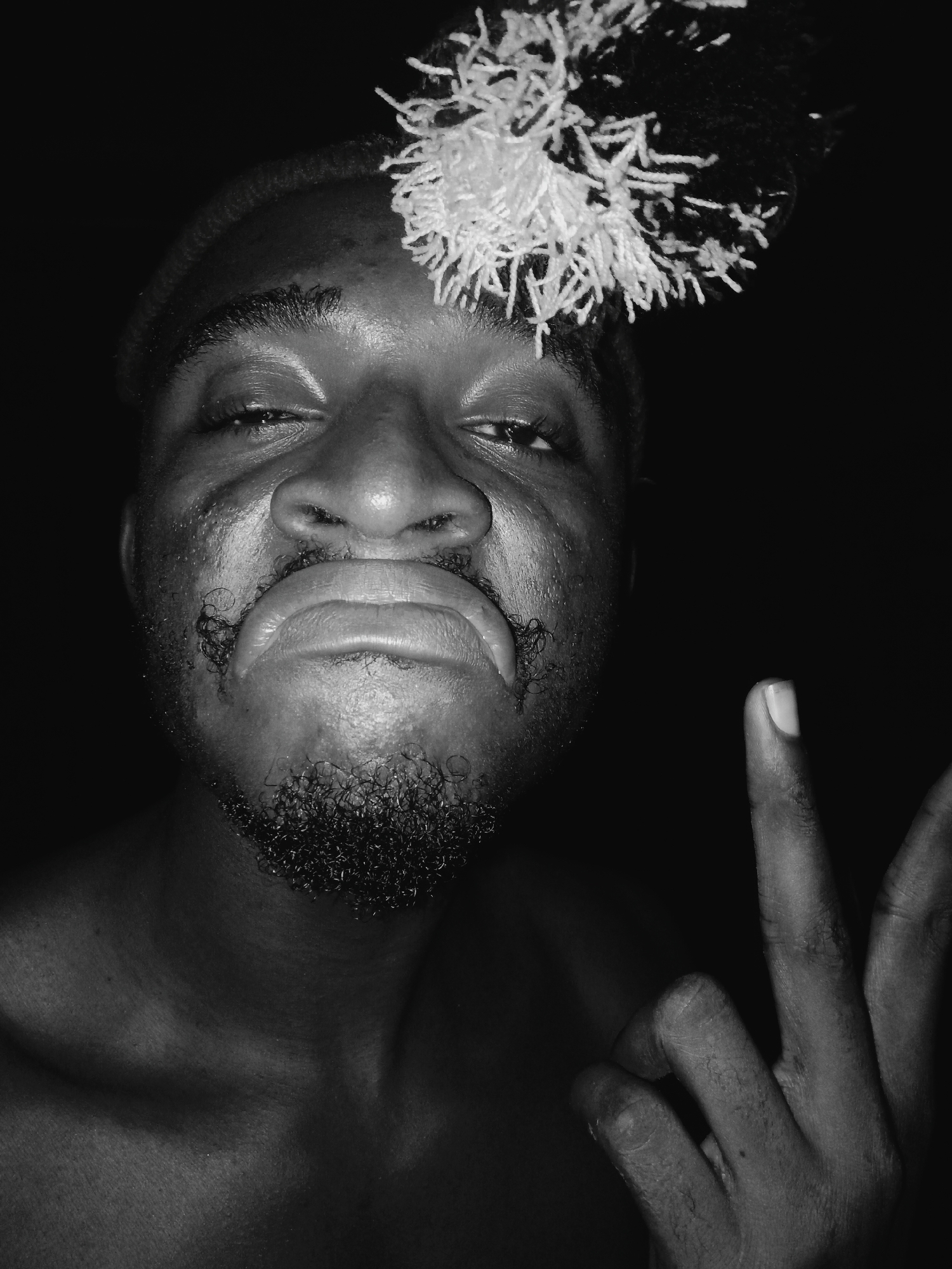 A close-up, black and white portrait captures a dark-skinned man, likely in Onitsha, Nigeria, against a completely dark background, suggesting a night-time or low-light indoor setting. The man's face, from his forehead to his upper chest, fills most of the frame. He wears a distinctive hat or head covering featuring prominent black and white yarn-like pom-poms or tassels on the top right side of his head.
His expression is exaggerated, with eyes narrowed and lips pursed into a pronounced pout, conveying a possibly playful, defiant, or self-assured emotion. He has a short, curly beard on his chin, and the hair around his upper lip is styled into small, distinct curls. The man's left hand (visible on the right side of the image) is raised, with his index, middle, and thumb fingers forming a gesture often interpreted as "OK" or the number three. The strong lighting highlights the textures of his skin, facial hair, and the details of his headwear. No text is visible in the image.