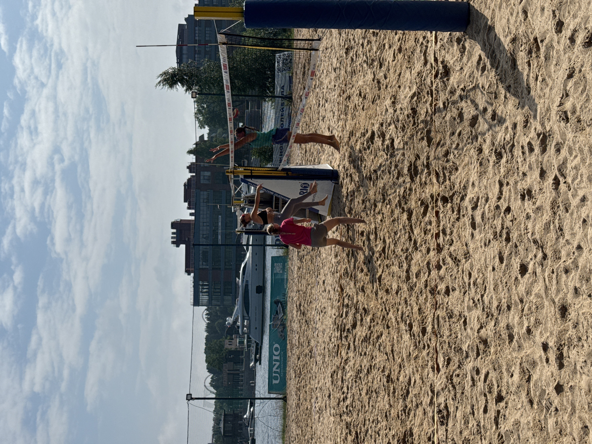 In this media file, two women are engaged in a beach volleyball game. The scene is a sandy beach volleyball court, with a net dividing the court. In the background, there are buildings, a body of water, a large yacht, and trees. The sky is visible with scattered clouds, suggesting it is daytime with fair weather.
One woman, wearing a teal tank top and blue shorts, is captured mid-air, diving to receive a ball. Her body is extended, and her arms are reaching out. The other woman, dressed in a pink top and gray shorts, is on the sand, also in motion, possibly preparing to play the ball.
Notable details include the textured sand of the court, the rigging of the volleyball net, and the sunlight casting shadows on the sand. The presence of the yacht and buildings in the background indicates an urban or coastal setting, consistent with the provided location context of Saint Petersburg, Russia. The image appears to be taken in the afternoon, given the angle of the shadows. The overall mood seems active and sporty, with the players focused on the game.
Visible text on a banner in the background includes "UNIO" and some Russian text, likely related to sponsorship or the event.