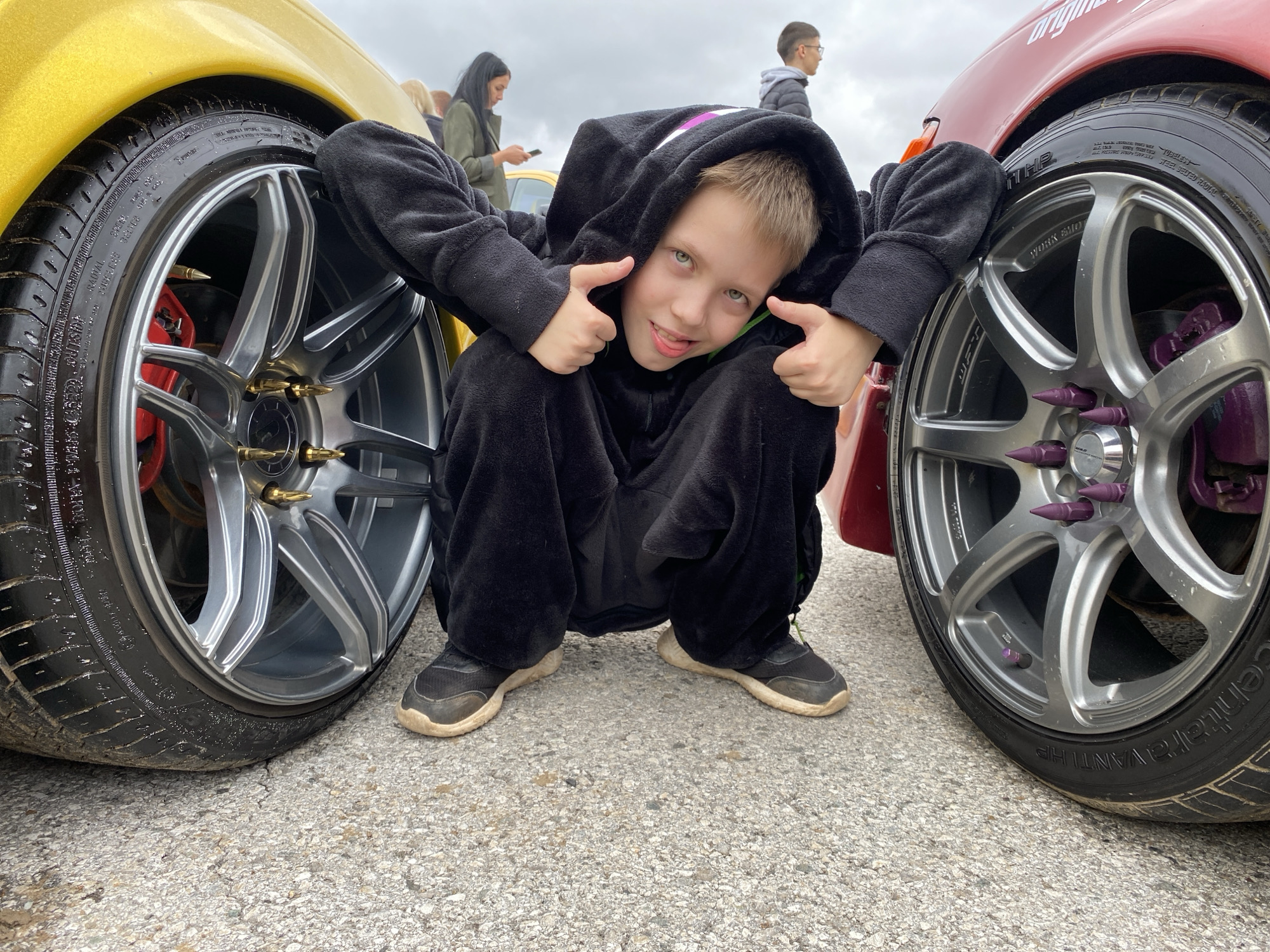 The image shows a young boy in a black onesie crouching between the front wheels of two cars, one yellow and one red. He is giving a thumbs-up with both hands. The cars have custom rims with purple and gold accents. The ground is asphalt with some debris. In the background, several other people are visible, some standing and looking at their phones. The sky is overcast. The image has a street photography feel, possibly taken at a car show or event. There is no indication of the location being the city of Perm, Russia.