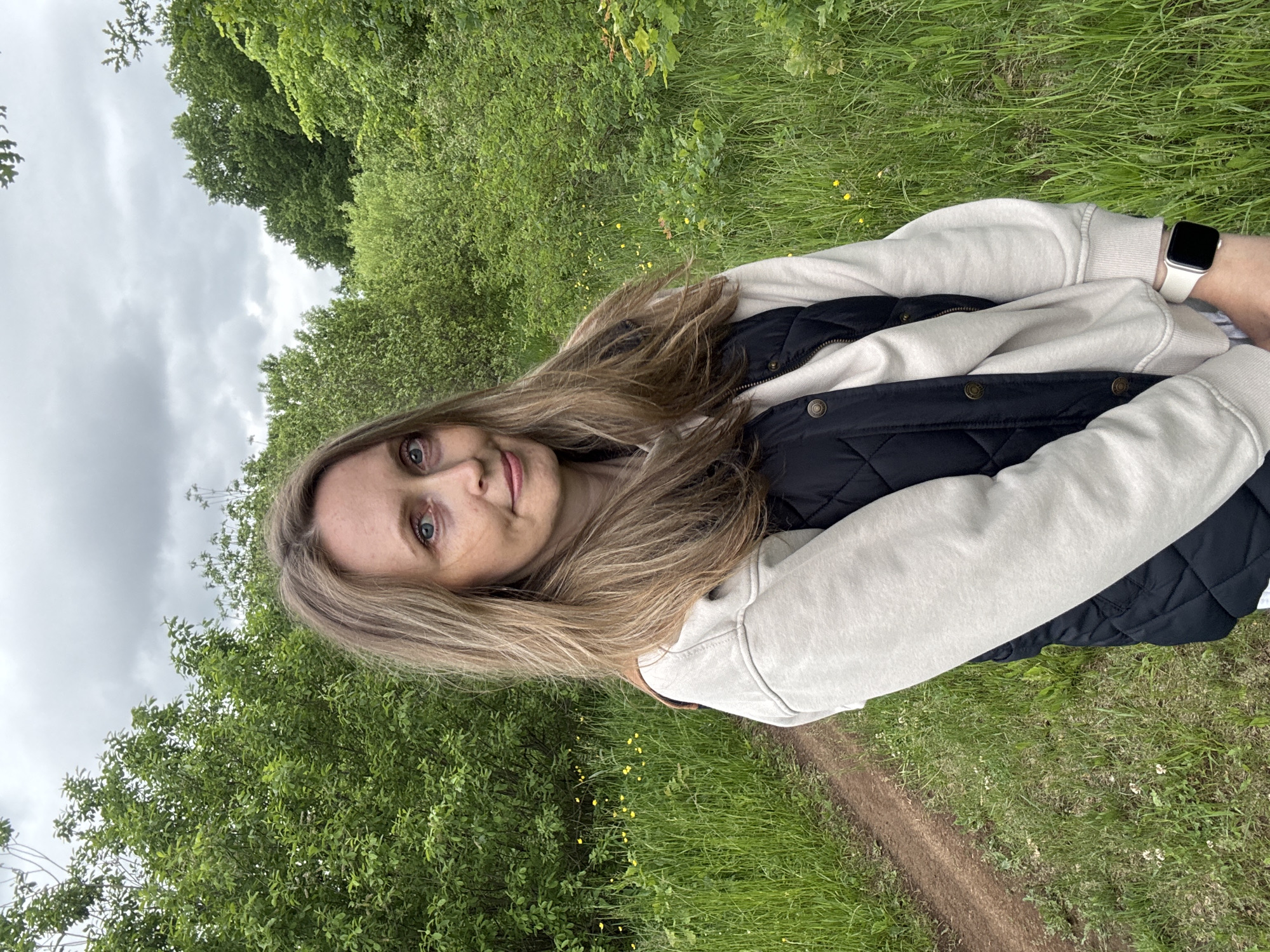 An adult female with long, light brown hair and blue eyes stands outdoors, facing forward with a faint smile. She is wearing an off-white long-sleeved top and a dark quilted vest with brass-colored snaps. A white smartwatch is visible on her left wrist. The environment is characterized by dense green foliage, including trees and shrubs, in the background and surrounding areas. To the left, a narrow dirt path winds through tall green grass dotted with small yellow flowers. The sky above is overcast with grey clouds. This scene is located in Noviy, Russia.