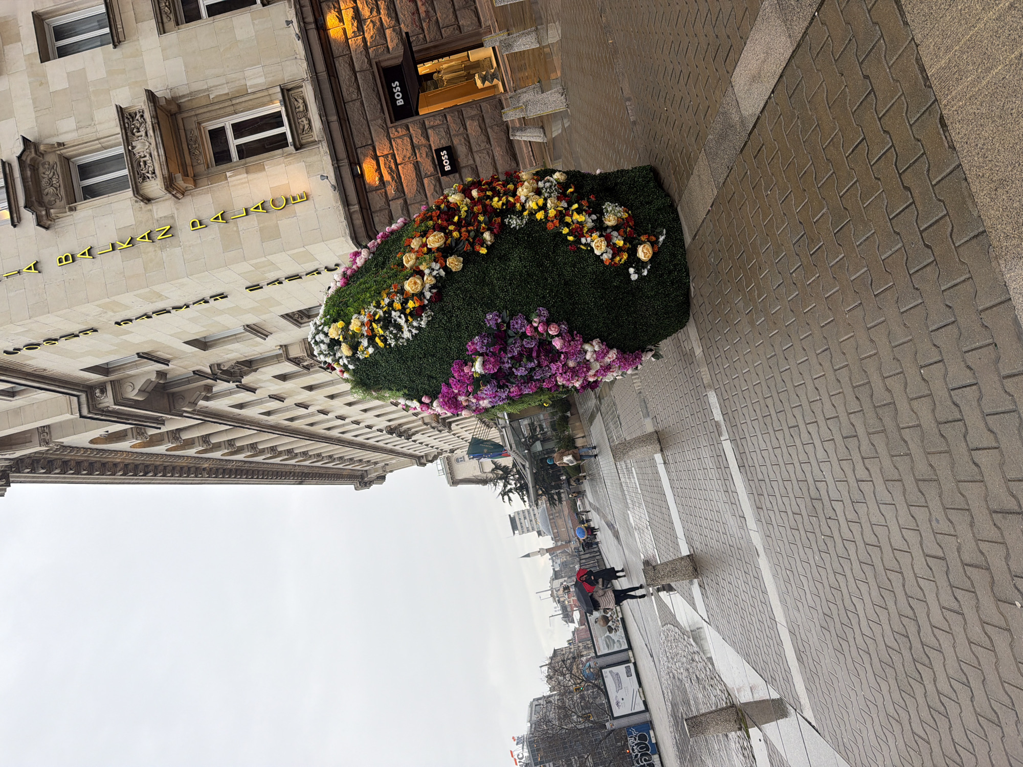 The image captures a street scene in Sofia, Bulgaria, likely along Vitosha Boulevard. A large, horizontal flower arrangement is prominent on the sidewalk in front of a building housing a "BOSS" store and the "Balkan Palace" Hotel. The flower arrangement features a variety of colors including white, yellow, orange, red, and purple. The sidewalk is paved with interlocking brick pavers. People are walking on the sidewalk in the distance, some with umbrellas. The sky is overcast.
