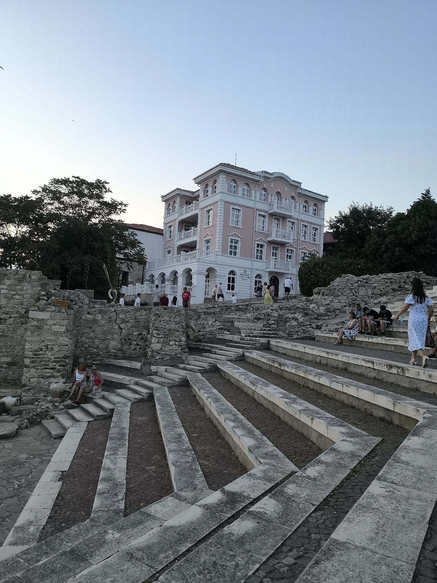 In Nesebar, Bulgaria, an outdoor ancient amphitheater and ruins are depicted. The ruins feature rough-hewn stone walls and tiered seating areas made of stone slabs. Several people are present, some sitting on the stone tiers and others walking. A prominent light pink building with white trim and balconies stands in the background, partially obscured by trees. The sky is a clear, pale blue, suggesting it is either early morning or late evening.