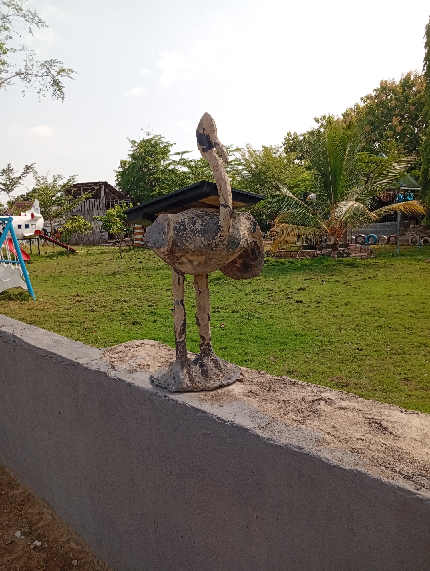 The image shows a concrete statue of an ostrich positioned on a concrete wall. In the background is a grassy field with trees and structures, including what appears to be a playground with a slide and airplane-shaped equipment. There is also a building with a thatched roof visible. The location is Adankolo, Nigeria.