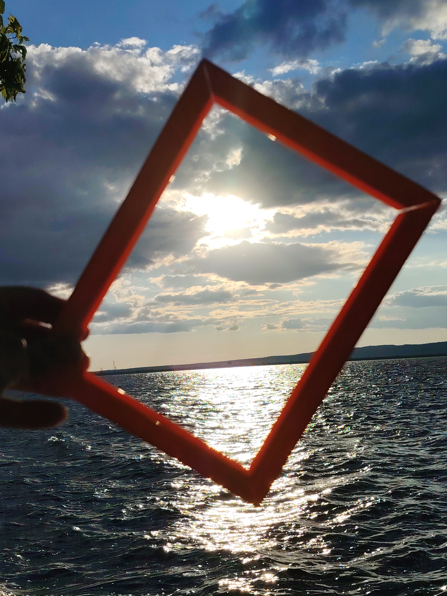 A first-person perspective shot shows a hand holding a bright orange square frame against a backdrop of a sky and a body of water. The sky features dramatic clouds and the sun breaking through, creating a bright glare on the water's surface. The water is dark and choppy, with sunlight reflecting off its waves. In the distance, a tree branch with green leaves is visible on the left side of the frame. The scene appears to be a natural outdoor setting, possibly overlooking a reservoir or large lake, with a distant shoreline and what looks like structures on the horizon. This specific view could be from the area of the Kama River in Chaikovsky, Russia, known for its scenic water bodies and natural landscapes.