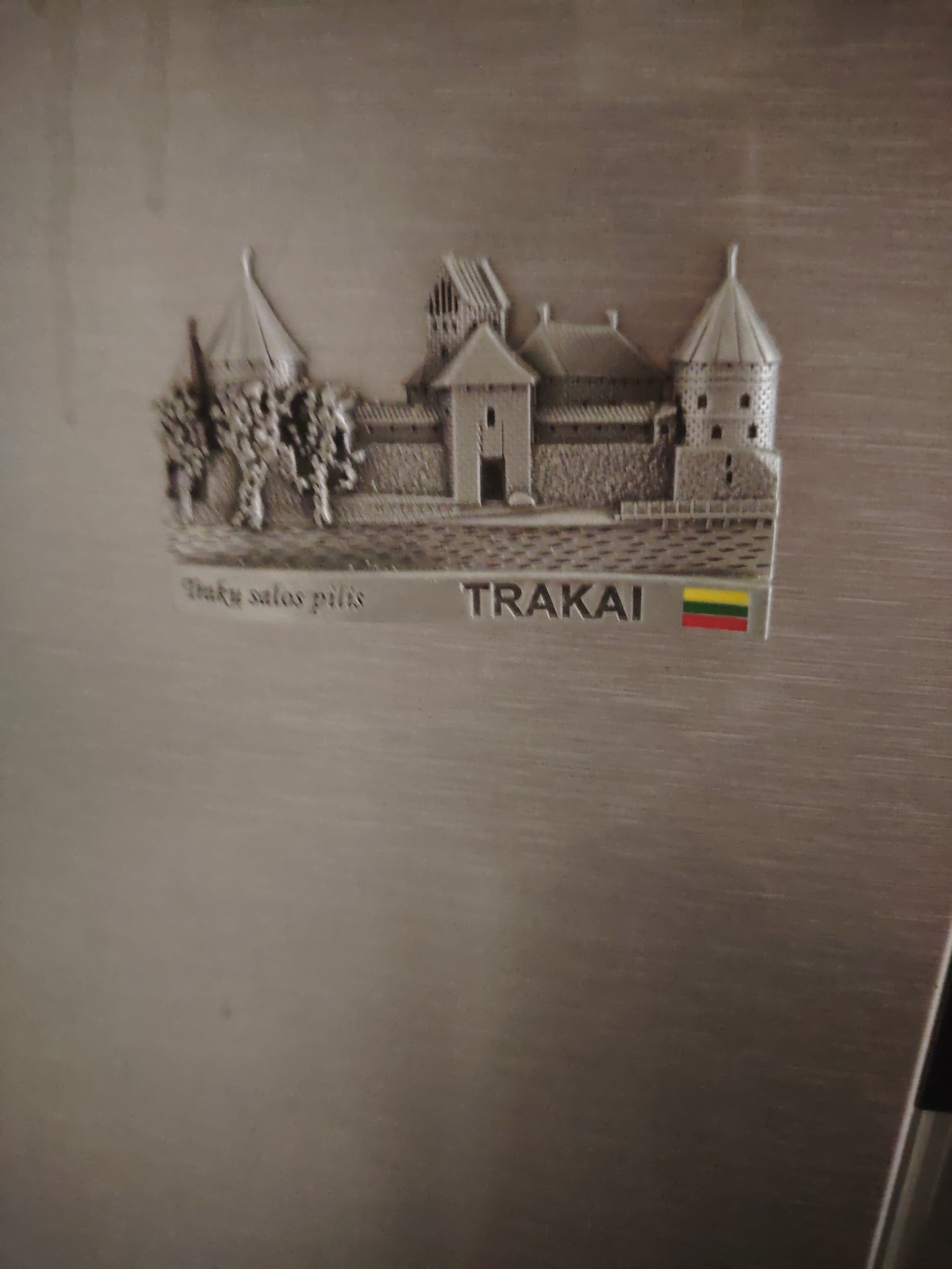 The image shows a silver refrigerator magnet shaped like a castle with the word "TRAKAI" and the Lithuanian flag printed on it. The magnet depicts a multi-towered castle structure with stone-like detailing and trees in the foreground. The text "Trakų salos pilis" is also visible, which translates to "Trakai Island Castle." The magnet is affixed to a brushed silver metallic surface, likely the door of a refrigerator. The background is out of focus and appears to be the interior of a room. There are no people or animals visible in the image. The location is not in Huntingdonshire, United Kingdom, as the magnet signifies Trakai, Lithuania.