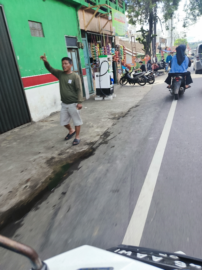 The scene is taken from the perspective of a person riding a motorcycle. The motorcycle is driving along a road in Ciwidey, Indonesia, located at the coordinates -7.103 latitude and 107.462 longitude. The road is paved with asphalt, and there is a white line down the center. On the right side of the road, a person wearing a blue jacket and a black headscarf is riding a motorcycle. The person is looking ahead and holding the handlebars of the motorcycle. The motorcycle has a black license plate and a black rearview mirror.
On the left side of the road, a man is standing near a gas station. He is wearing a green shirt, gray shorts, and black sandals. The man is waving his hand in the air, as though he is trying to flag down a vehicle. The man is standing in front of the gas station, which has a white sign with the word "Pertamina" on it. The gas station has two pumps. The man has a phone in his hand, but it is not clear whether he is using it. Behind the man is a green building with a red stripe on the bottom. The gas station is located on the sidewalk, which is made of concrete.
The scene is taken on a sunny day. The sky is blue, and the trees in the background are green. There are other vehicles, including a white car and a motorbike, parked on the road.