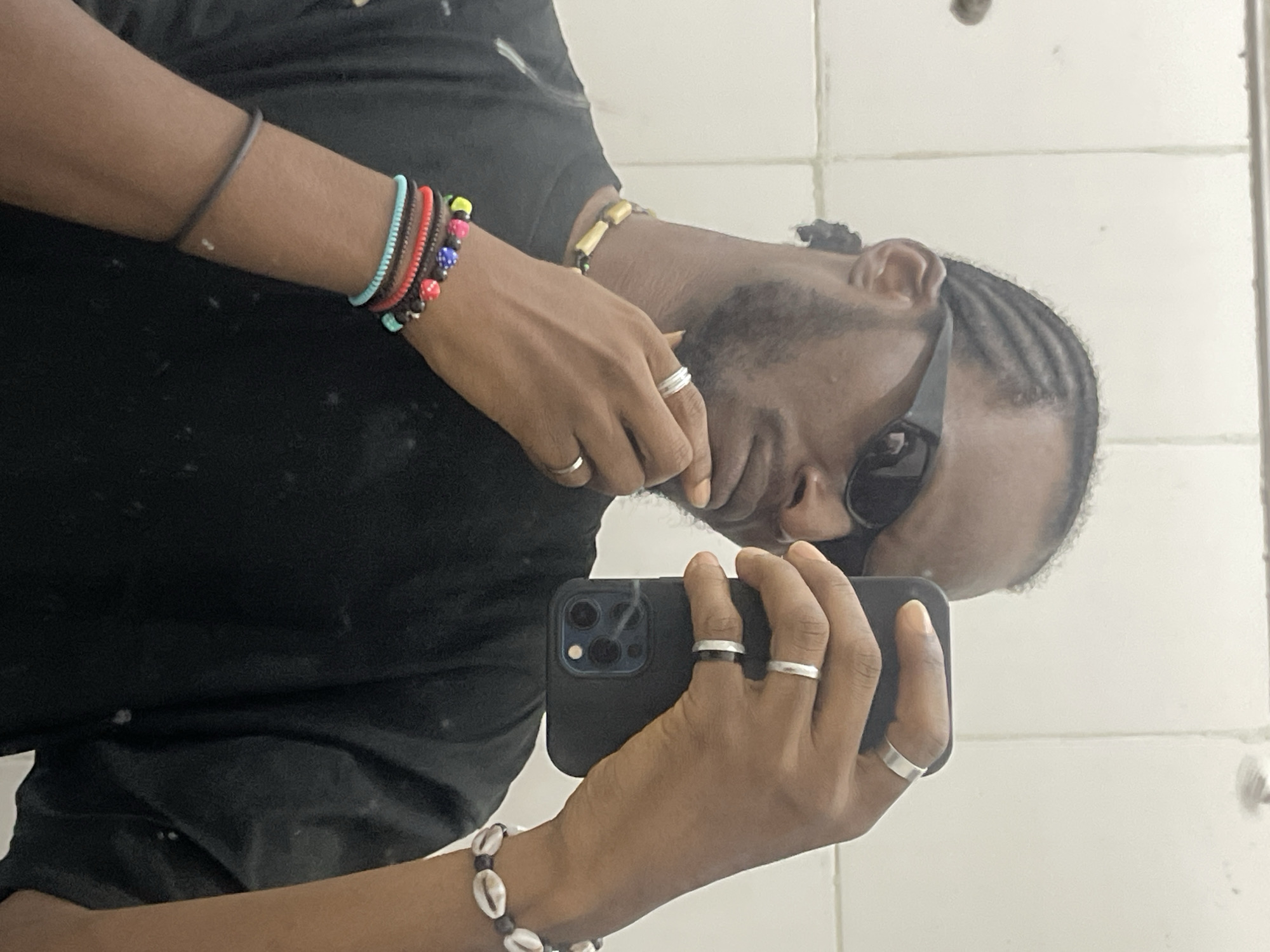 A male individual with dark skin captures a mirror selfie in what appears to be an indoor setting with white tiled walls, located in Jos, Nigeria. He wears a black short-sleeved t-shirt, black rectangular sunglasses, and has a neatly trimmed beard and mustache. His hair is styled in cornrow braids pulled back into a small tie at the nape of his neck.
His left hand is raised, with the fingers lightly touching his chin. On his left hand, he wears a wide, matte silver-toned band on his ring finger and a thinner, matte silver-toned band on his pinky finger. His left wrist is adorned with multiple bracelets: a colorful beaded bracelet (featuring pink, blue, yellow, and red beads), a dark red coiled or wrapped bracelet, a turquoise coiled or wrapped bracelet, and a single thin dark band further up his forearm. He also wears a beaded necklace with light-colored, cylindrical beads and smaller dark beads.
His right hand holds a black iPhone, likely a Pro model given the visible triple camera array on the back, positioned to take the mirror photograph. On his right hand, he wears a wide, matte silver-toned band on his index finger and thinner, matte silver-toned bands on his middle and ring fingers. His right wrist features a bracelet composed of white cowrie shells interspersed with dark beads.
The background consists of light-colored, possibly white or off-white, rectangular tiled walls with visible grout lines. The tiles and mirror surface show minor smudges or dust particles.