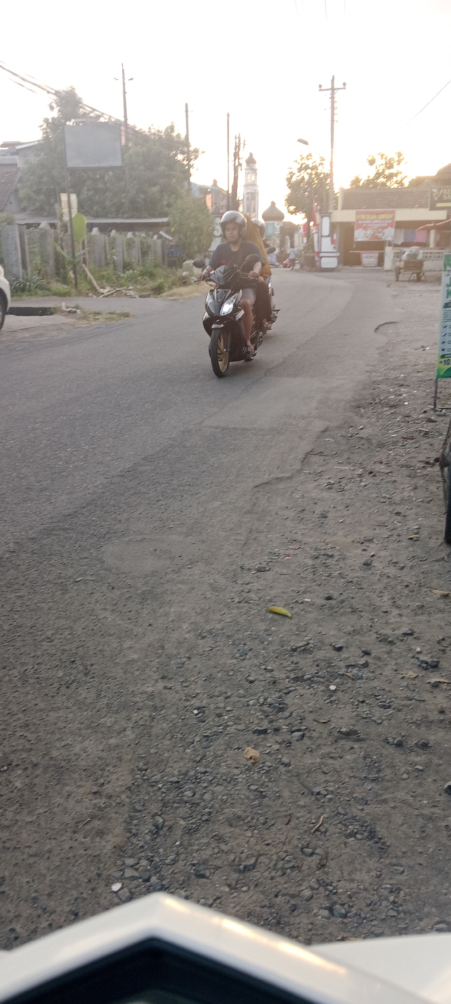 A man wearing a black helmet rides a black motorcycle with a passenger behind him on a paved road in Maguwoharjo, Indonesia. The man is wearing a dark shirt and shorts. The passenger is wearing a yellow top. The road is lined with low fences, trees, and buildings. A tall white minaret and dome structure, likely a mosque, is visible in the background, along with utility poles and power lines. The sky is bright and appears to be either early morning or late afternoon, with the sun casting a warm glow. The road surface shows signs of wear and is covered with gravel and some debris. In the foreground, the edge of a white vehicle is visible.