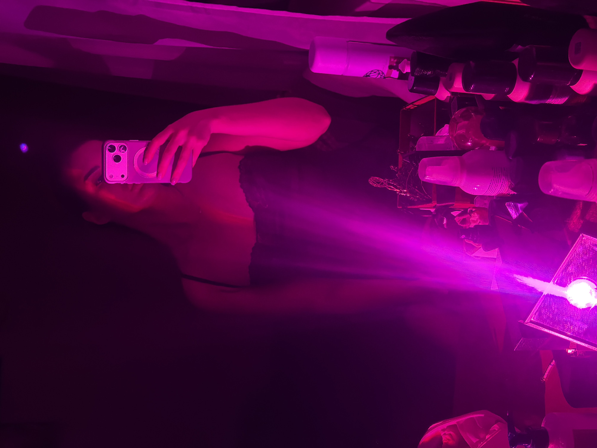 The image shows a person holding a pink smartphone and taking a selfie. They are standing in a room with dim pink lighting. Behind the person is a shelf with various items including bottles and containers. There is a light fixture or lamp emitting a bright beam of pink light. The location and city, Moscow, Russia are not evident from the image.