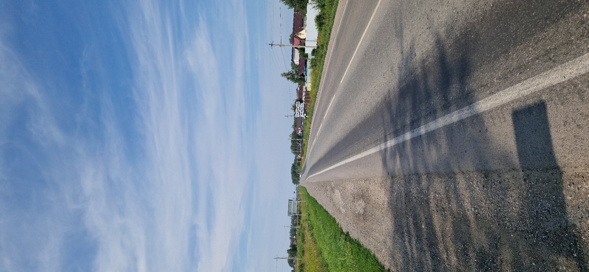 The image depicts a wide, paved asphalt road in the countryside, likely near the city of Leninskoye, Russia. The road has a white painted line marking the lanes and a gravel shoulder on the left side. To the right of the road, there are several houses with red tiled roofs and fences, suggesting a residential area or village. Beyond the houses, trees and vegetation are visible, and further in the distance, more trees line the horizon. The sky is blue with scattered white, wispy clouds. There are power lines and utility poles along the side of the road, and a few vehicles can be seen in the distance on the road. A sign with text is visible near the right side of the road, though the text is not legible in detail. The overall impression is of a sunny day in a rural or semi-rural setting.
