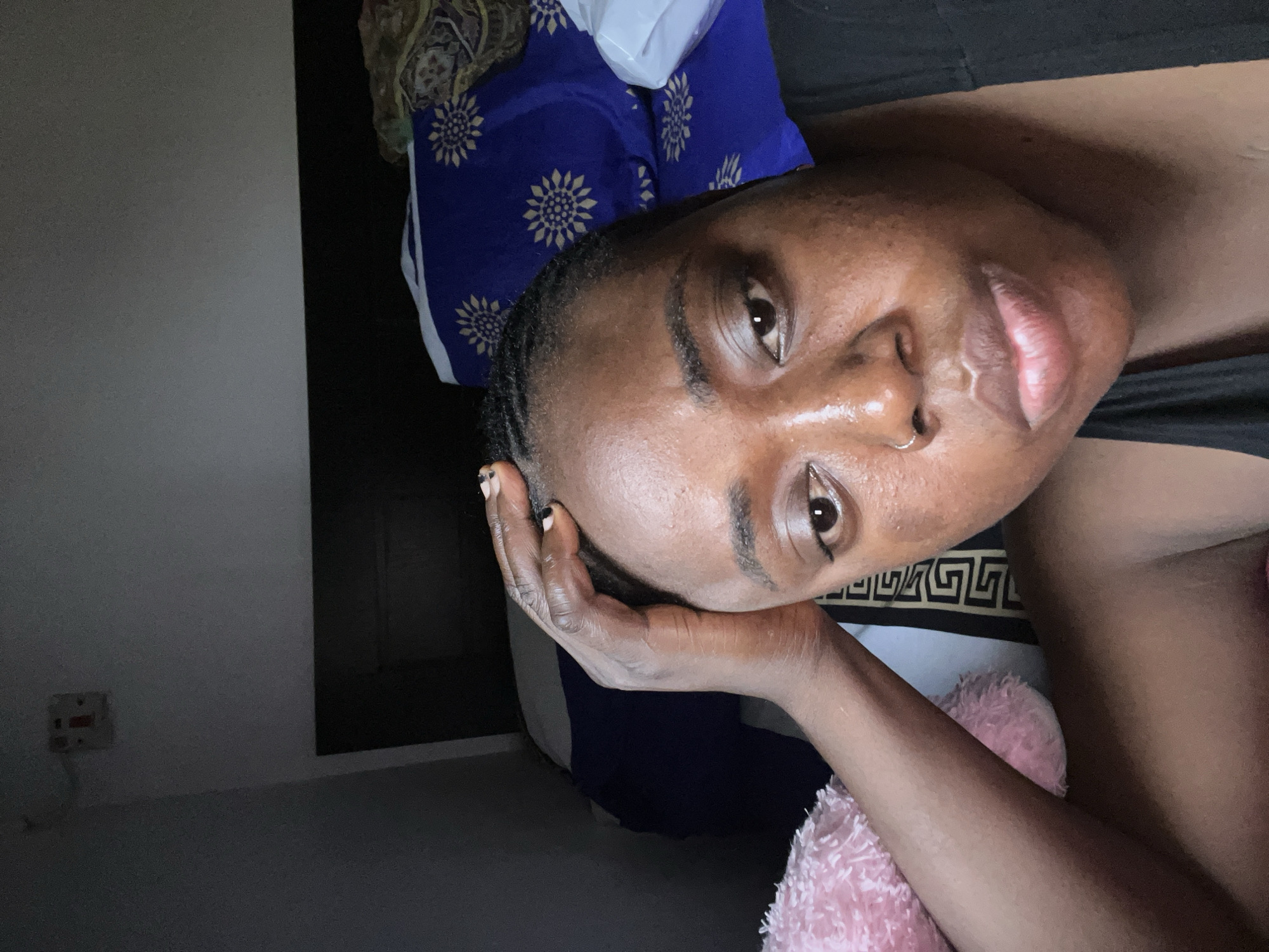 A medium shot captures a woman lying down, looking directly at the camera in a relaxed indoor setting. She has dark skin, brown eyes, and dark eyebrows, with a small nose ring adorning her right nostril. Her dark hair is styled close to her head. Her left hand, featuring dark nail polish with lighter tips, supports her head under her chin, contributing to a calm and contemplative expression. She is wearing a dark, possibly black, top.
The background elements suggest a bedroom setting in Abuja, Nigeria. To the left, a plain light grey wall is visible, with a partially obscured electrical outlet. Behind her head is a vibrant blue fabric, likely a pillowcase or bedding, patterned with repeating golden sunburst or floral designs. A dark area to the right might be a headboard or furniture. Underneath her right arm and shoulder, a fluffy pink blanket or throw is visible, alongside a black fabric with a gold/white Greek key pattern, presumably part of the bedding. The dim lighting suggests it is evening or nighttime, creating a soft, intimate atmosphere. There is no visible text.