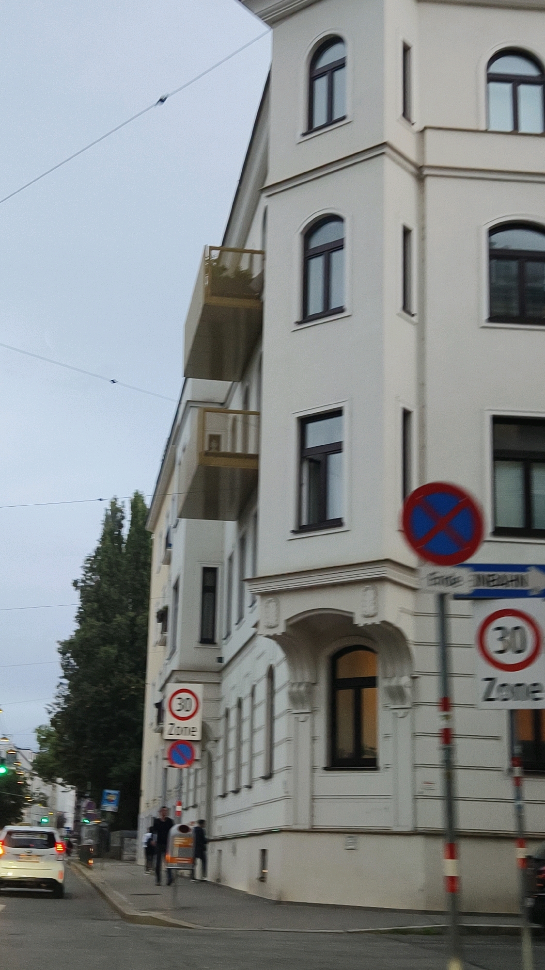 The image depicts a street scene in Vienna, Austria, featuring a white apartment building with several windows and a balcony. On the right side of the building, a traffic sign with a red circle and a blue cross, indicating no parking, is visible above a sign for a "30 Zone." Further to the left, another "30 Zone" sign is present. A white car is driving on the street in the foreground, and two individuals are walking on the sidewalk. A tall, green tree stands to the left of the building. The sky is overcast.