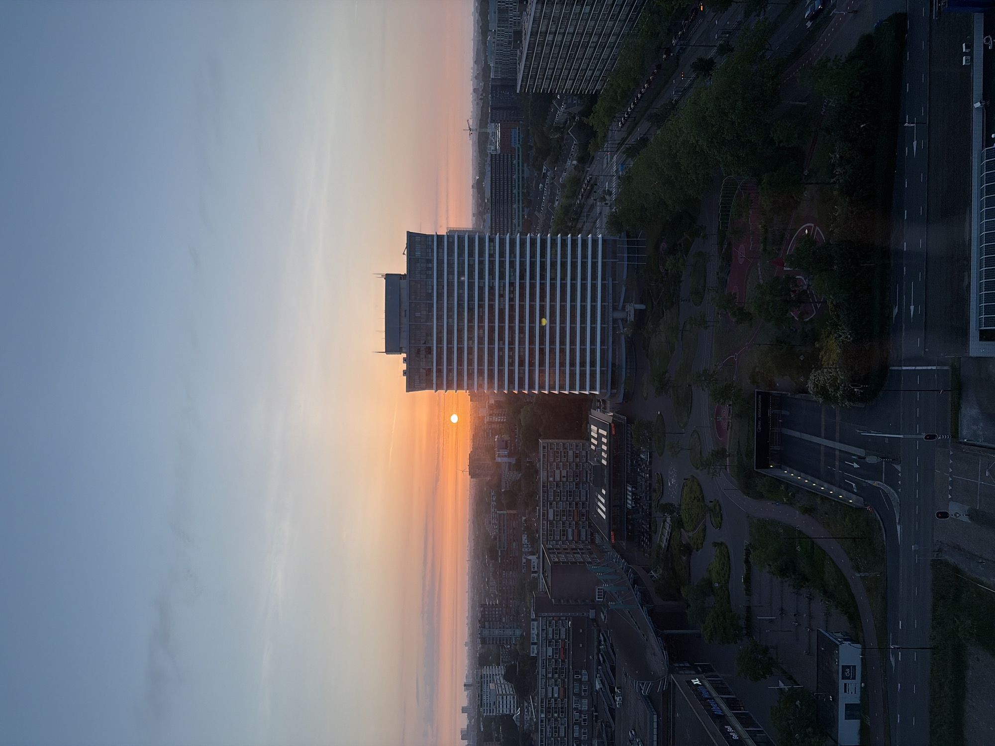 An aerial view of Rijswijk, Netherlands, at dawn or dusk. The sky transitions from pale blue to orange. The sun is low on the horizon, obscured by a tall building with horizontal lines across its facade. Below the building, there are dense buildings, and farther in the distance, the city extends under a muted orange sky. To the right of the tall building, a park with a running track is present, adjacent to a road and what seems to be a tunnel. The environment shows an urban setting at a time of day where the light is soft and limited.