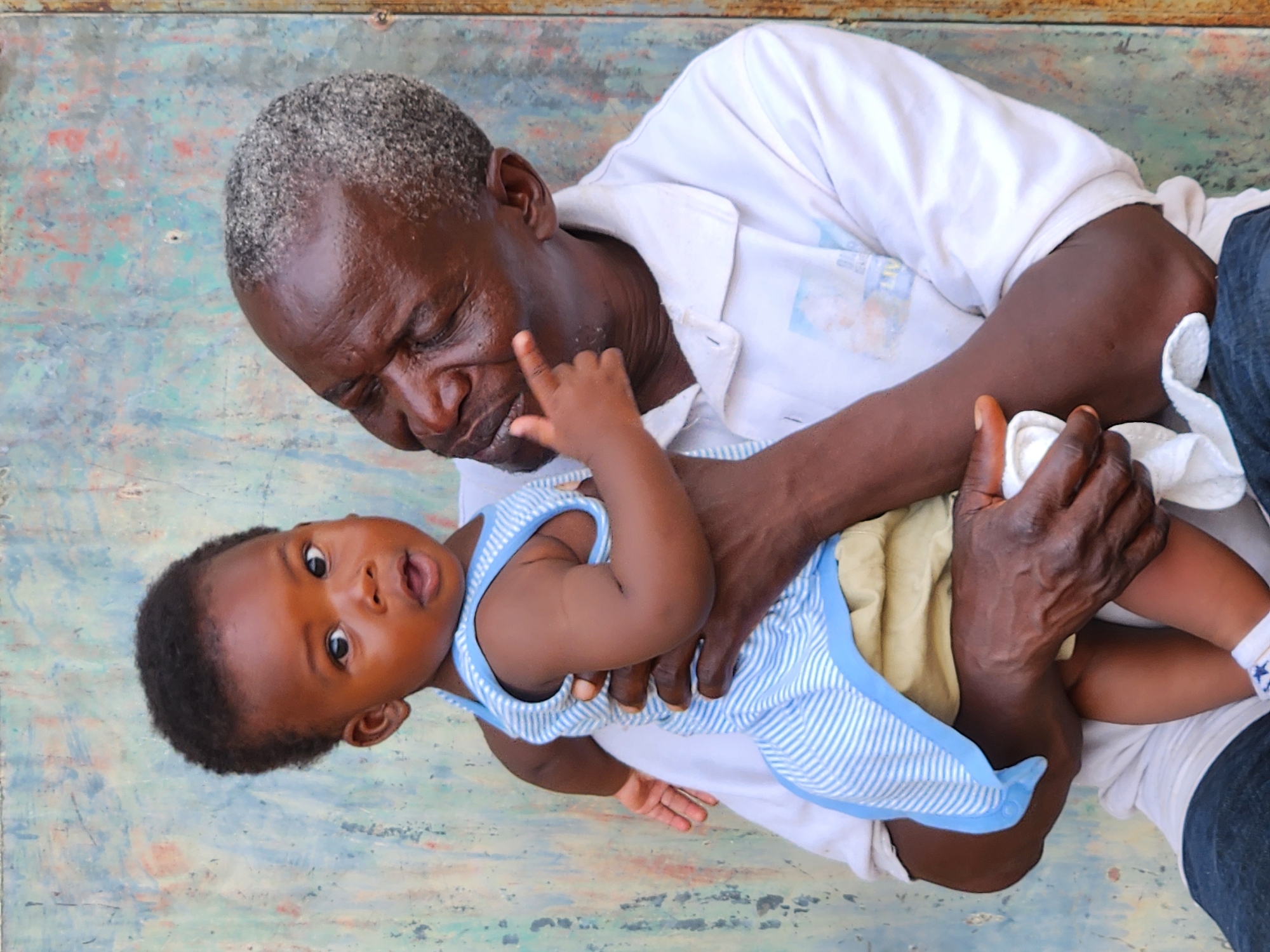 An elderly Black man with graying hair is holding a baby in his arms against a weathered, teal and rust-colored wall. The man is wearing a white collared shirt with a faded image on the front. The baby is wearing a light blue and white striped onesie with a pale yellow bottom. The baby's left hand is reaching up towards the man's face, with a finger touching his cheek. The man's right arm is around the baby. The scene appears to be outdoors, possibly in a residential area, given the state of the wall. The image is a medium shot, focusing on the interaction between the man and the baby. The setting suggests a warm climate.