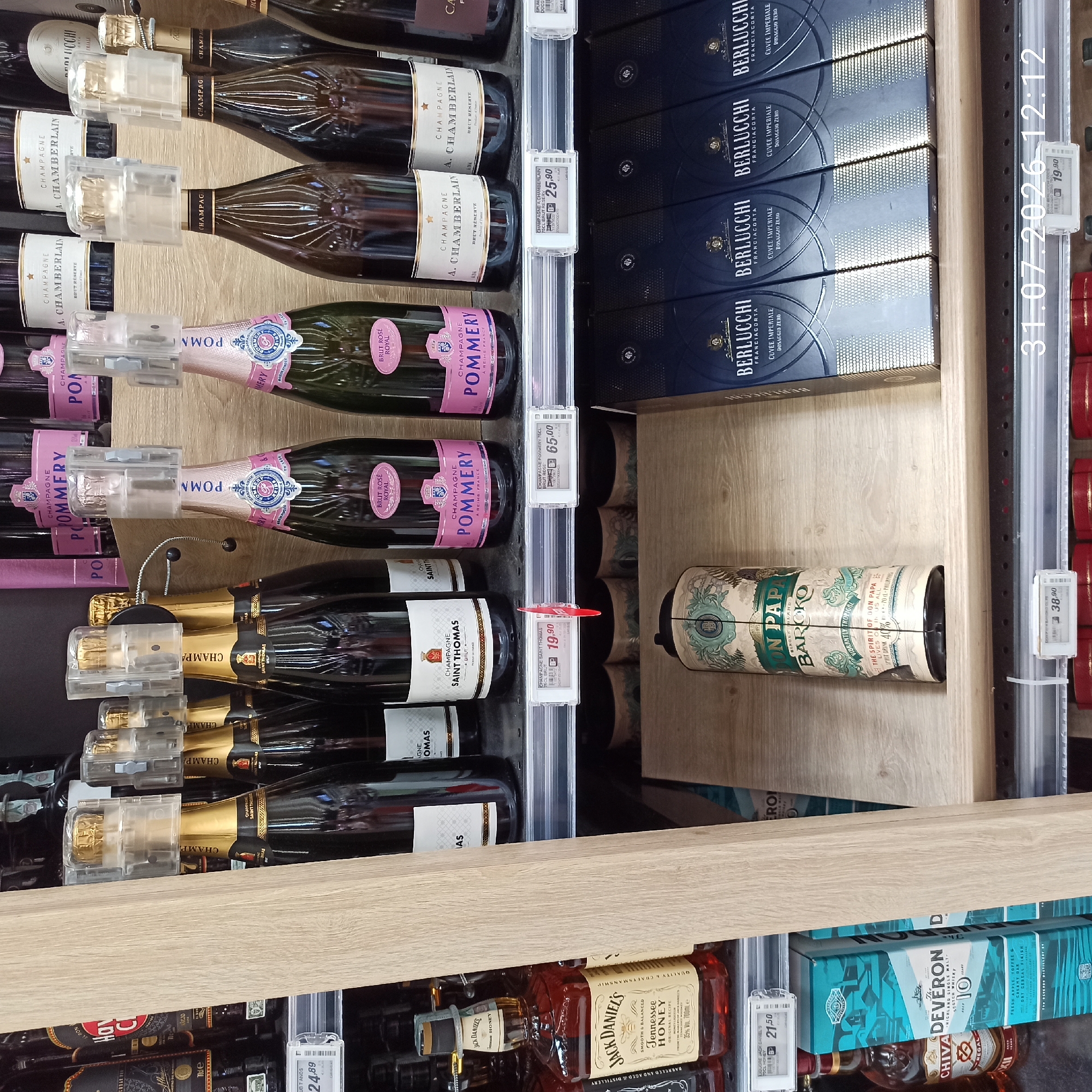The image shows a retail store aisle with several shelves displaying alcoholic beverages. On the top shelf, there are bottles of champagne, including "POMMERY" and "CHAMPAGNE SAINT THOMAS". On the middle shelf, there is a bottle of Jack Daniel's Tennessee Honey and several bottles of champagne. Below that, on another shelf, there is a decorative bottle of "DON PAPA BAROKO" and several boxed bottles of "BERLUCCHI FRANCIACORTA". The lighting appears to be artificial, typical of a retail environment. The date and time stamp on the bottom right of the image reads "31.07.2026 12:12". The image is a photograph taken within the city of Modugno, Italy, likely in a supermarket or liquor store.