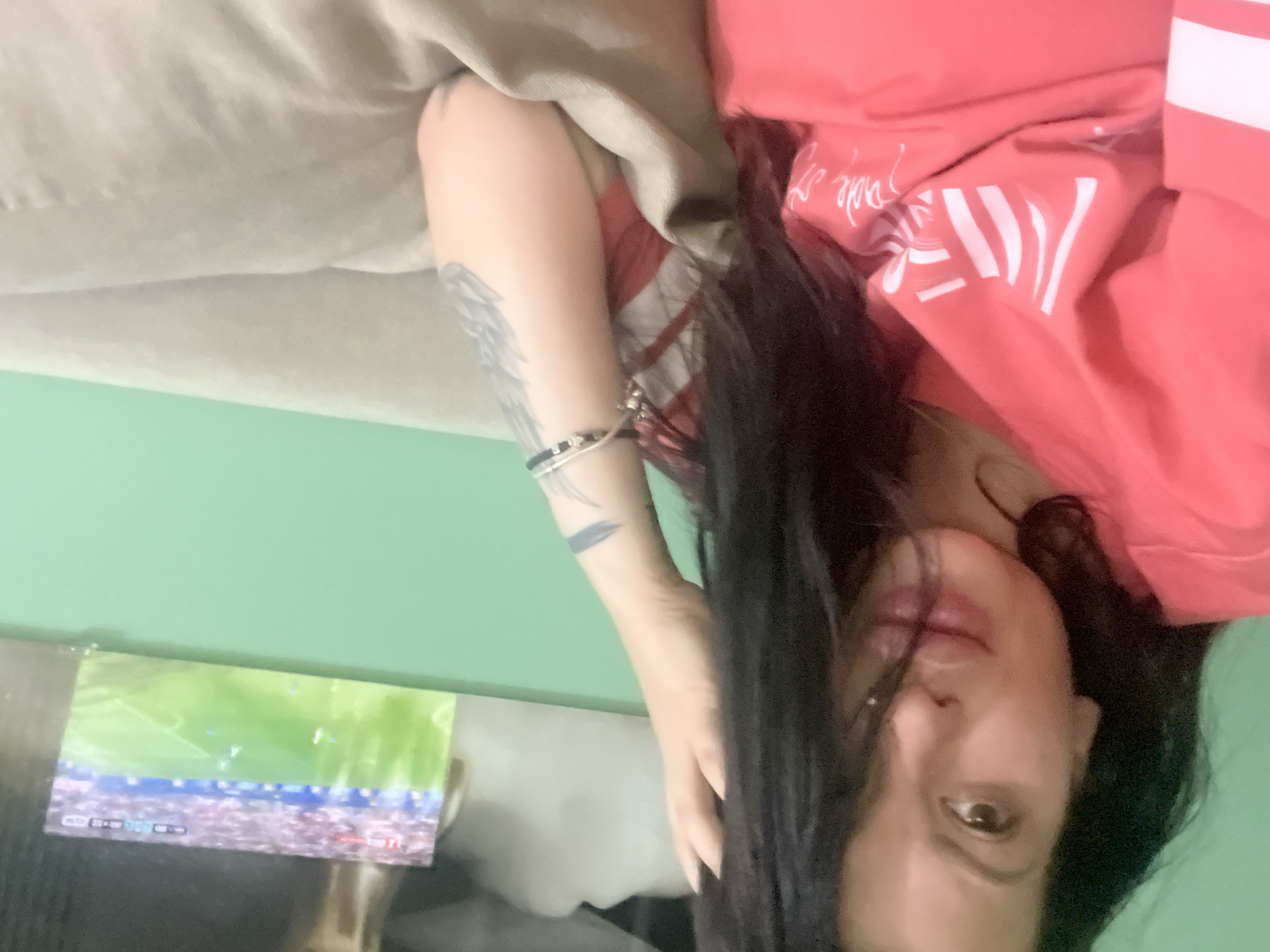 In a close-up, indoor shot, a woman with dark hair is seen from the chest up, resting on a light-colored sofa. She is wearing a pink t-shirt with white stripes and lettering. Her left hand is placed on her head, and her right arm rests on the sofa. She has tattoos on her right arm and is wearing bracelets. In the background, a television screen displays a soccer match. The wall behind the sofa is painted light green. The scene appears to be a living room. There is no information in the image to suggest the location is Plovdiv, Bulgaria.