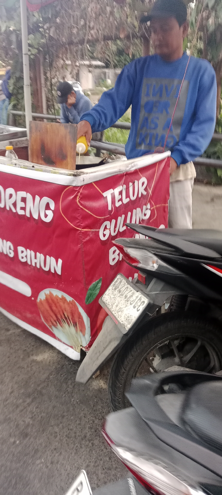 This is a close-up, eye-level, medium shot of a food cart with a red banner in what appears to be an outdoor urban setting in Ngemplak, Indonesia. A person wearing a blue sweatshirt and a black baseball cap is standing behind the cart, holding a yellow squeeze bottle and appears to be dispensing a liquid into a cooking pan. The red banner on the cart has white text that reads "ORENG", "BIHUN", "TELUR", and "GULUNG". To the right of the banner, a portion of a black motorcycle is visible, including its rear wheel, fender, and license plate. The license plate is partially obscured and difficult to read, but some characters are visible. The background is blurred and shows foliage and possibly some structures, indicating an outdoor environment. The overall scene suggests a street food vendor in operation.