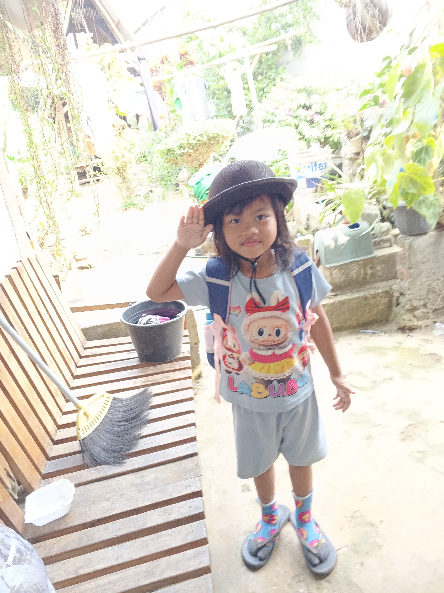 A young girl wearing a black hat, a light blue t-shirt with cartoon characters and the word "LABUBU" on it, and light blue shorts stands outdoors in a slightly blurry image. She has a backpack with pink straps. The girl is raising her right hand to her forehead as if saluting or waving. She is wearing colorful socks and flip-flops. To her left, a wooden bench is visible with a black bucket filled with clothes and a broom with a yellow handle lying on it. In the background, there is lush greenery, a building with a tin roof, and some concrete steps. The environment appears to be a residential backyard or courtyard, likely in Warungkiara, Indonesia, given the tropical vegetation and outdoor setting.