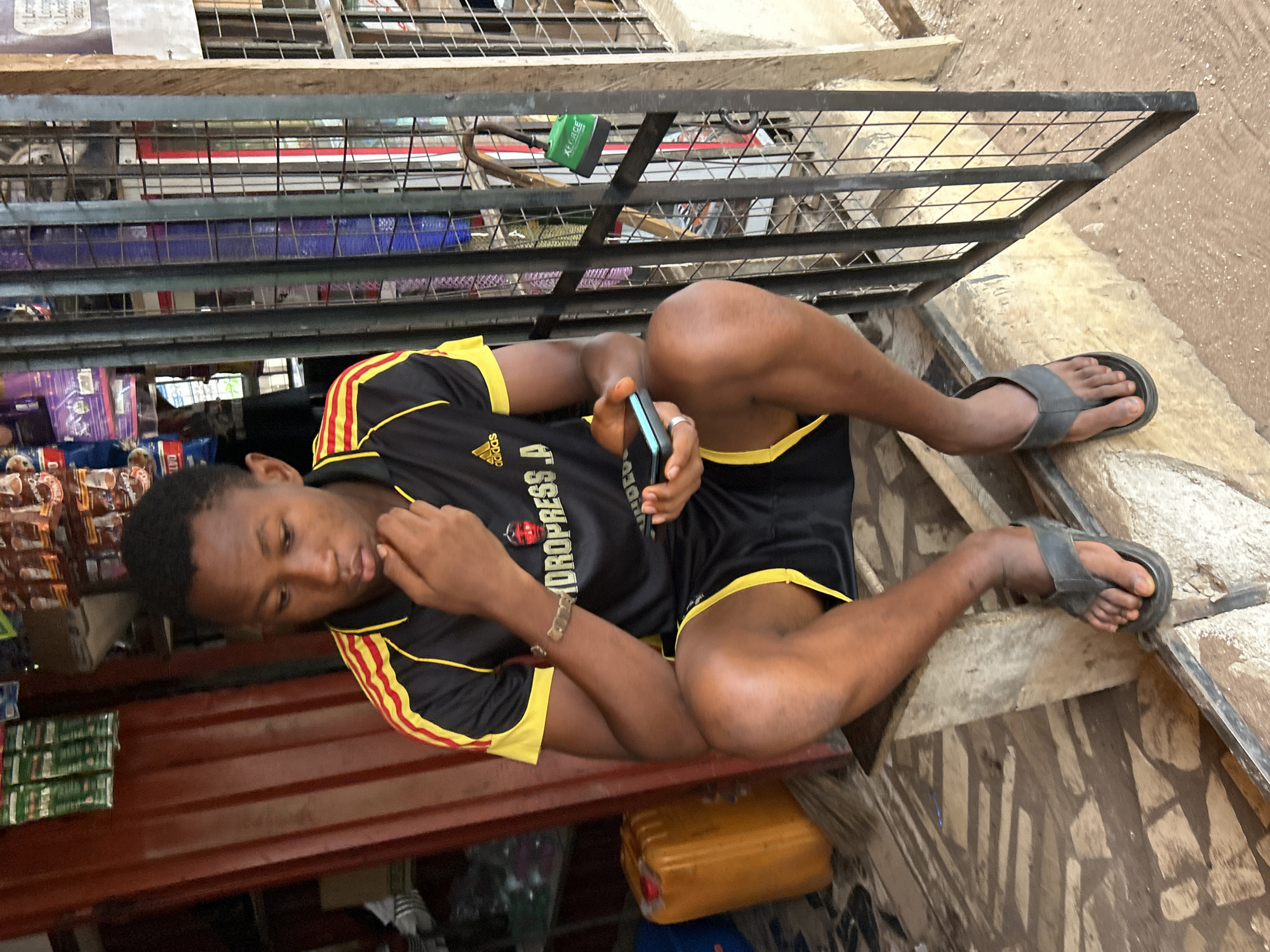 A young Black male, appearing to be in his late teens or early twenties, is seated on a concrete step outside a small shop. He is wearing a black short-sleeved t-shirt with yellow and red accents, and black shorts with yellow trim. His right hand is resting on his chin, and he is looking down at a smartphone he holds in his left hand. He is also wearing a silver-colored bracelet on his left wrist and grey flip-flops on his feet.
The setting appears to be an outdoor market stall in Ihiagwa, Nigeria. Behind the young man, on the left, is a red corrugated metal structure, likely part of the shop's exterior, with various packaged goods visible on shelves within. To the right, a metal grate encloses the storefront, with items displayed behind it. A bright yellow plastic container is visible on the ground to the left of the young man. The ground surface is a mix of sand and what appears to be patterned concrete or stone. A green padlock is attached to the metal grate. The overall lighting suggests it is daytime.