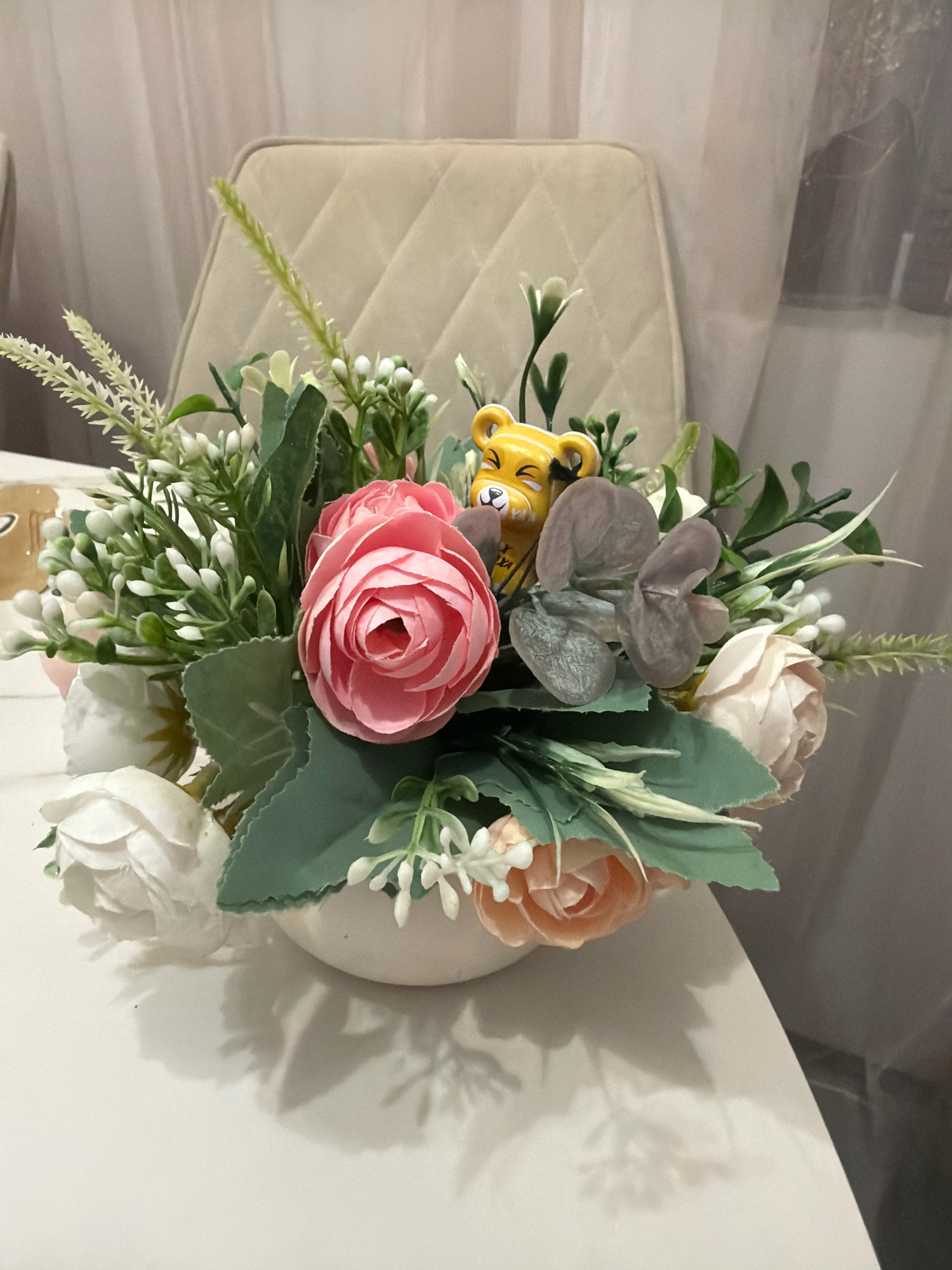 An artificial floral arrangement sits on a white surface in what appears to be an indoor setting. The arrangement is composed of various flowers, including pink roses, white roses, and peach-colored roses, interspersed with greenery and small white buds. A small, yellow toy tiger figurine is nestled among the flowers. In the background, a quilted beige chair and sheer curtains are visible. The scene is illuminated, casting shadows of the arrangement onto the white surface. The location of Vniissok, Russia, is not evident from the visual information in the image.