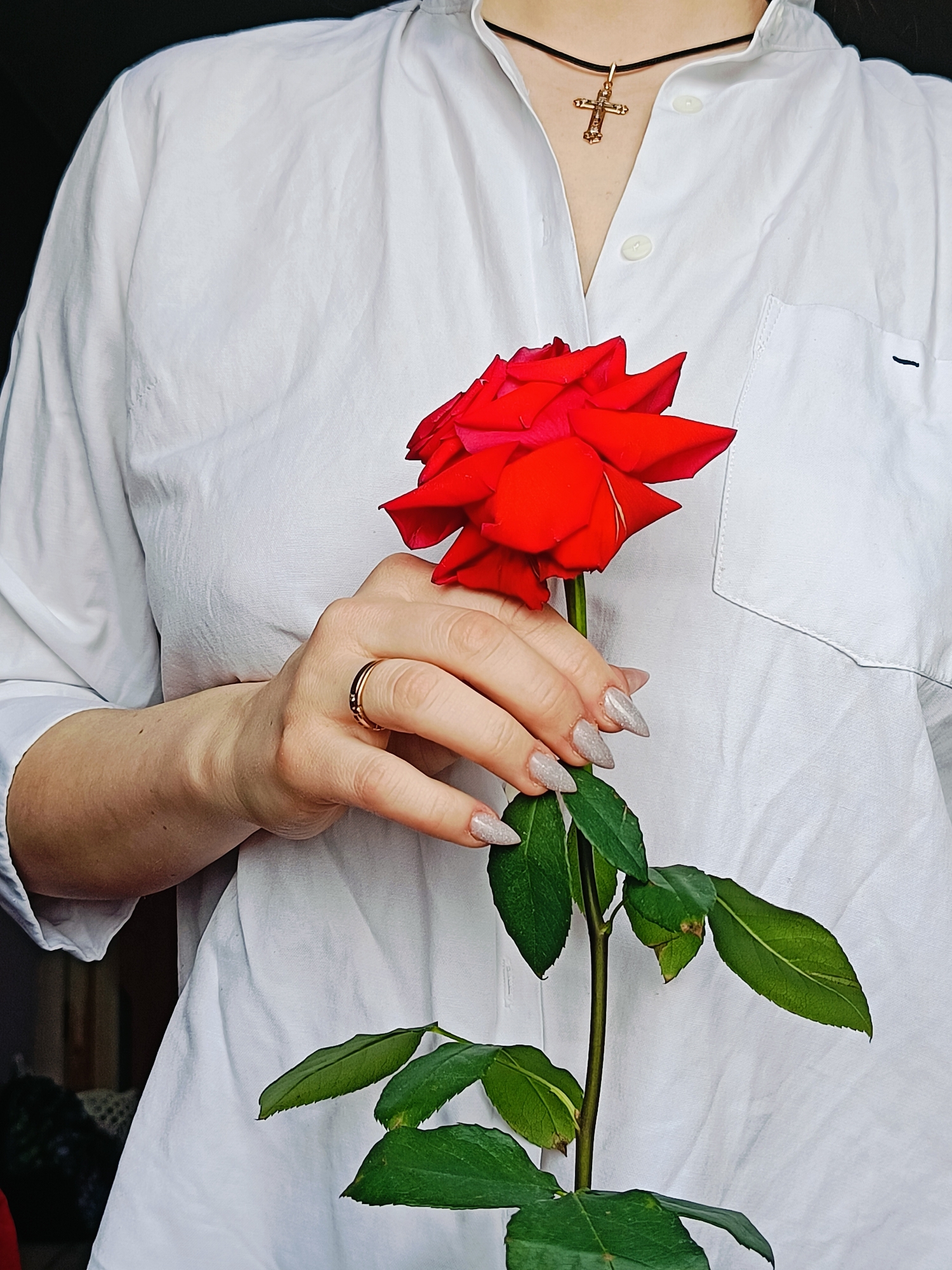 A person is holding a red rose. The person is wearing a white button-down shirt, a black choker with a gold cross pendant, and a gold ring. The rose has green leaves and a long stem. The person's fingernails are long and decorated with a light-colored glitter polish. The background is out of focus, but it appears to be indoors. No information is available on whether this picture was taken in Rusalevka, Russia.