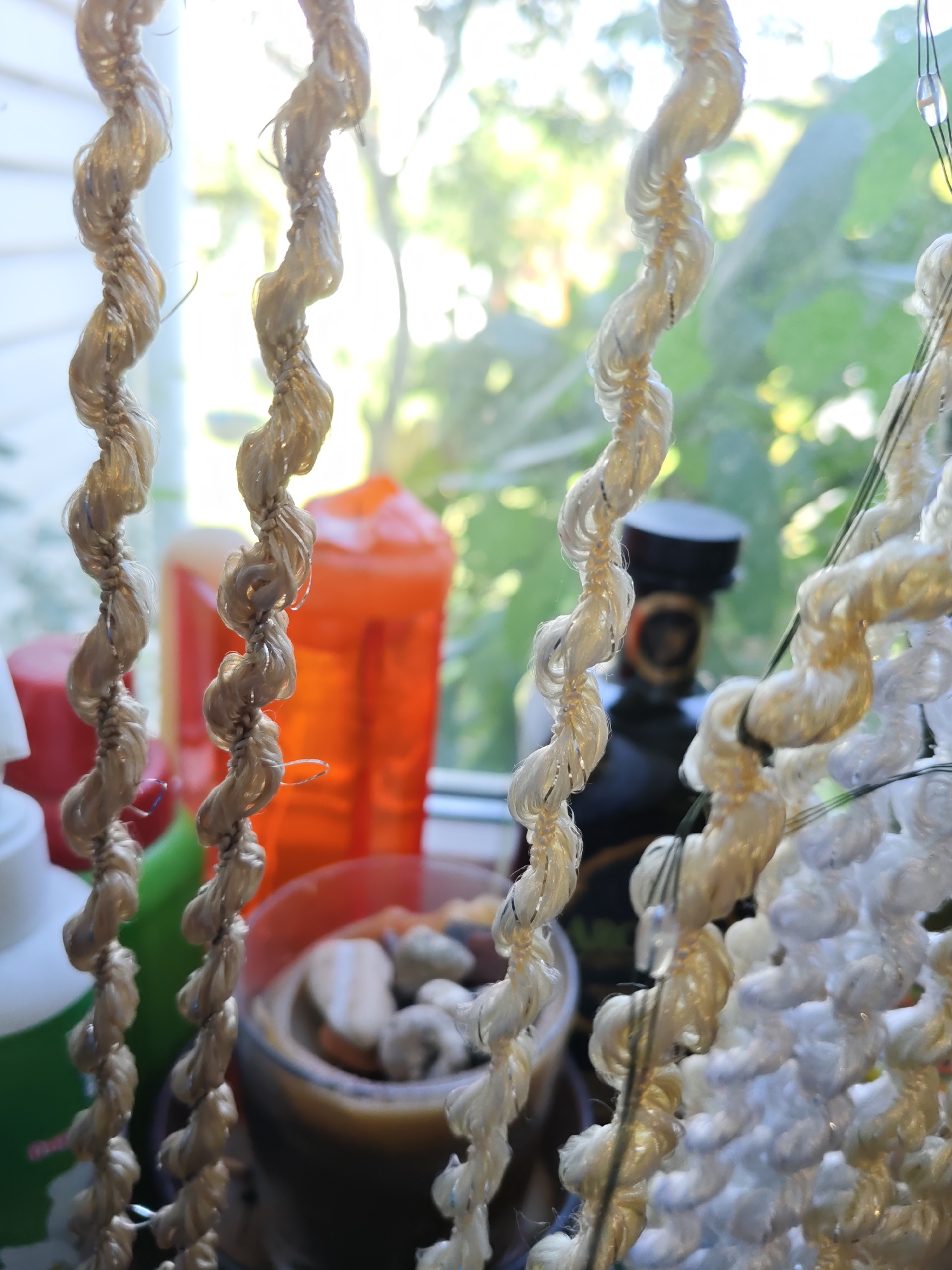 The image is a close-up, shallow depth of field shot of several twisted strands of light-colored, possibly jute or sisal, rope hanging vertically in front of a sunlit window. The rope has a textured, organic appearance. Behind the rope, the blurred background suggests an indoor setting with a window that lets in natural light, illuminating green foliage and potentially a building exterior. On a surface beneath the window, various objects are visible but out of focus, including an orange plastic container, a dark bottle with a black cap, and a cylindrical object that might be a candle in a glass holder containing stones. The overall impression is one of a still life, with the focus on the textural details of the hanging ropes. The location of Rostov-on-Don, Russia is not evident from the image.