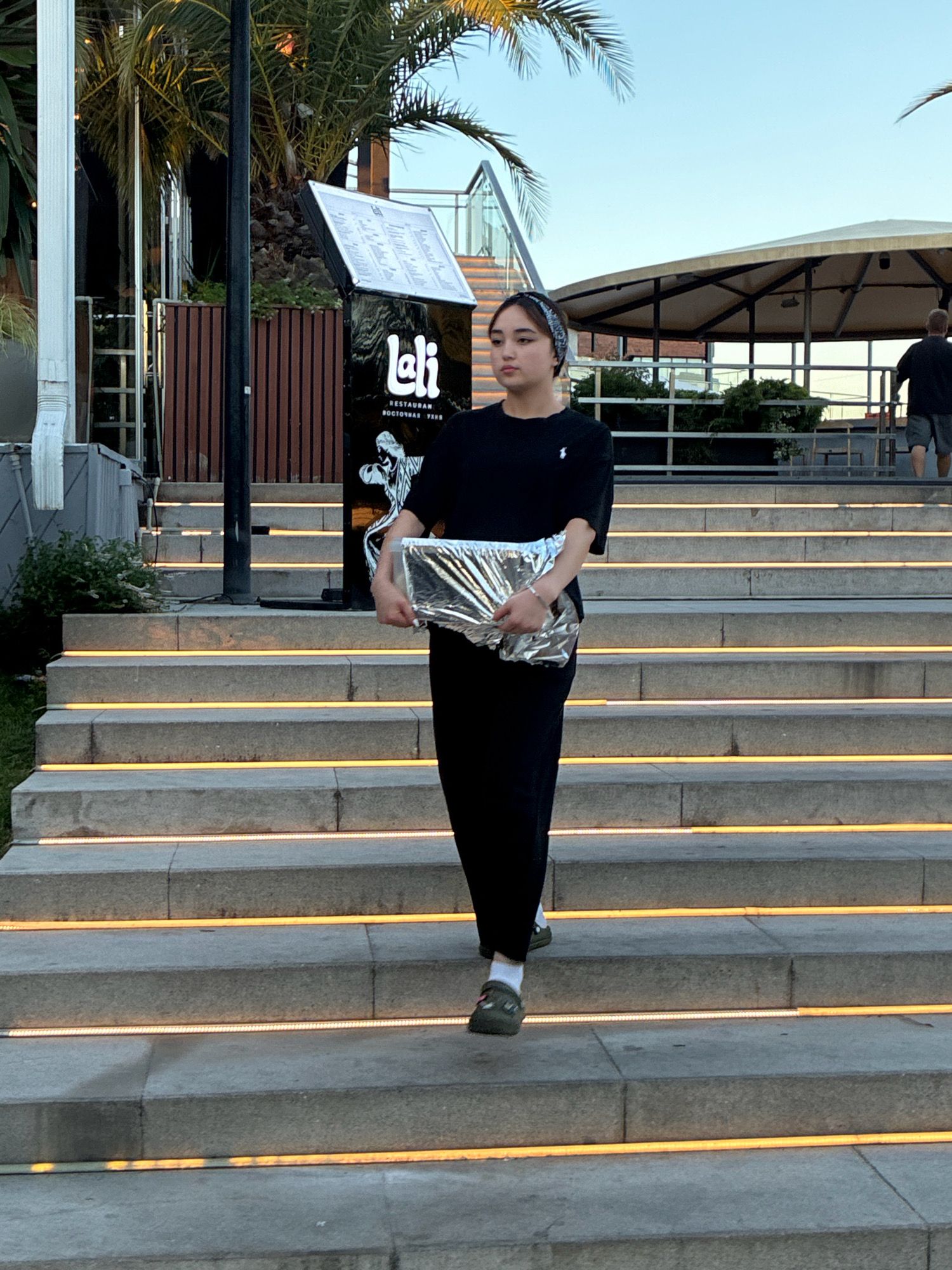 A young woman with dark hair tied back with a patterned bandana walks down a wide, outdoor staircase at an illuminated venue in Sochi, Russia. She is wearing a black t-shirt with a small white logo, black pants, and green slip-on shoes with white socks. She carries a shiny, crinkled silver bag in her right arm. The staircase is made of stone with bright orange LED lights embedded along the edges of each step. Behind her, a sign for "lali" restaurant is visible, with palm trees and outdoor seating structures in the background. The sky is clear and blue, suggesting it is daytime or early evening. Another person, a man in a dark shirt and shorts, is visible walking up the stairs in the distance. The overall scene depicts a modern, outdoor dining or entertainment area.
