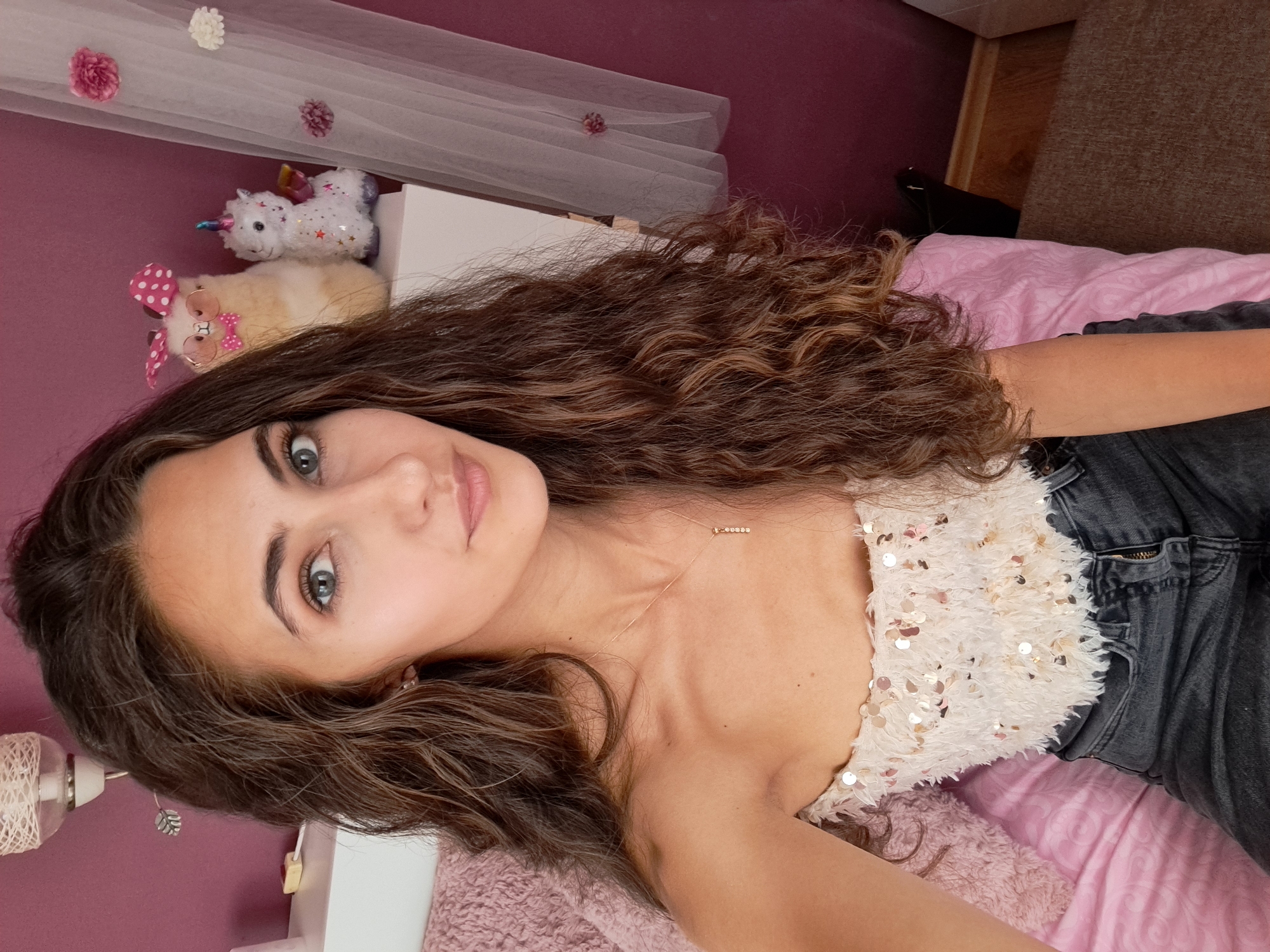The image displays a head-and-torso portrait of a female subject, positioned centrally, looking directly at the camera. The individual has long, wavy, dark brown hair, fair skin, grey-blue eyes, and is wearing a light-colored, strapless top embellished with shimmering sequins and a textured, possibly feathery, surface. Dark grey denim jeans with a visible zipper fly are also worn. A delicate gold-colored necklace with a small, vertical pendant featuring five small, embedded elements adorns her neck. Small, circular, light-colored earrings are visible.
The subject is seated or reclining on a bed with pink bedding, including a textured pink blanket on the left and a smoother pink sheet or duvet on the right. The background features a deep magenta-colored wall. A white headboard or shelf extends horizontally behind the subject. On the left side of the headboard, a wall-mounted light fixture with a woven white shade is visible, along with a small yellow object on the white surface. On the right side of the headboard, two plush toys are displayed: a light brown llama-like figure wearing a pink polka-dot bow tie, matching head wrap, and round glasses, and a white unicorn figure with a rainbow horn, star embellishments, and small wings. To the far right, a sheer white fabric, possibly a canopy or curtain, is visible, decorated with several artificial flowers (pink and white). The lower right corner reveals a light brown carpet or rug next to a dark object. The scene is illuminated by indoor lighting.
Location: Unknown, Unknown.