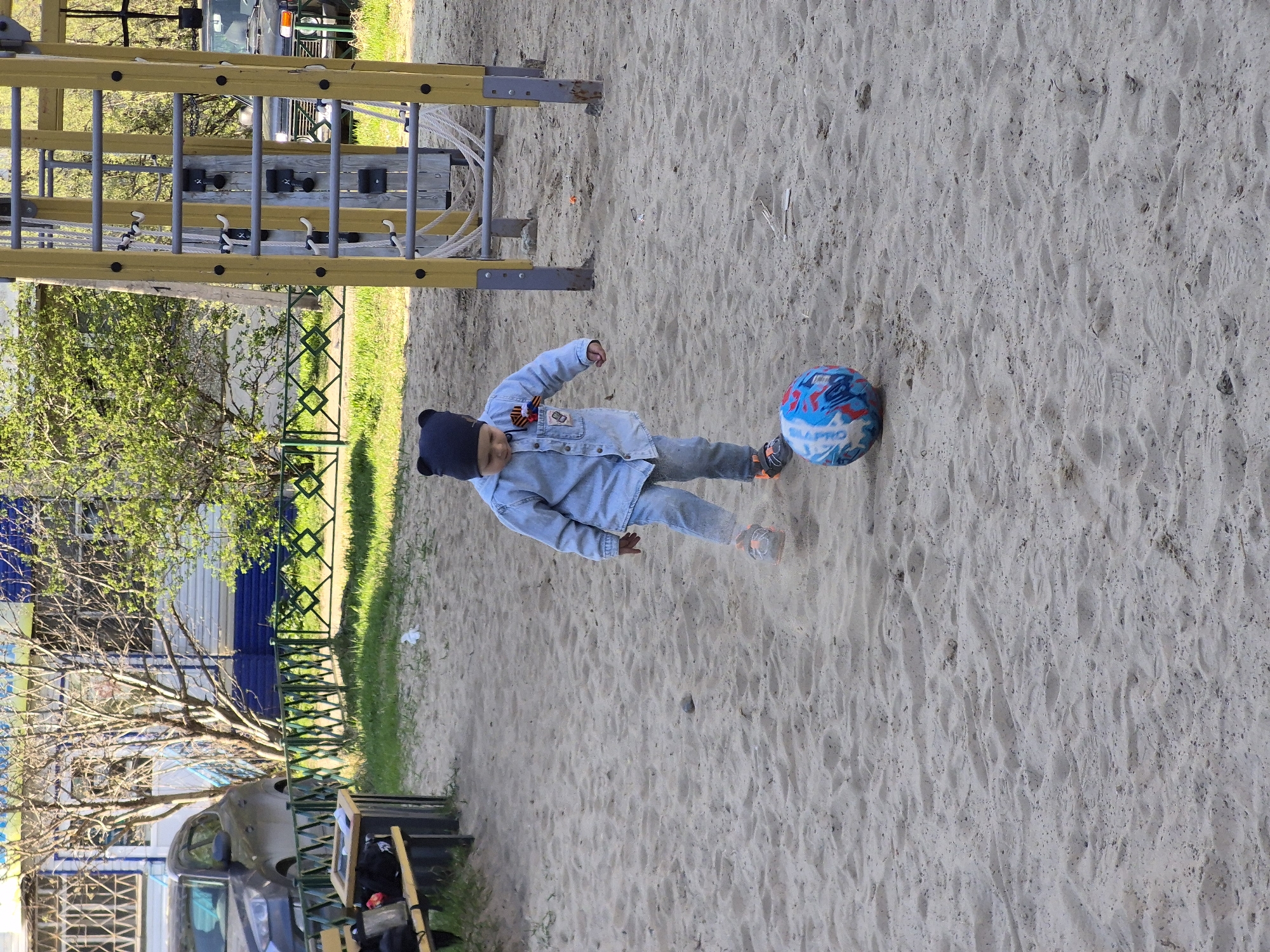 A young child is kicking a blue and white ball in a sand-covered playground. The child is wearing a dark blue hat with bear ears, a light blue denim jacket, and light blue jeans. The playground features a climbing frame with yellow and silver bars in the upper left corner. In the background, there is a green fence, a tree with sparse foliage, and parked vehicles including a van. The specific location in the city of Tobolsk, Russia, cannot be determined from the given image.