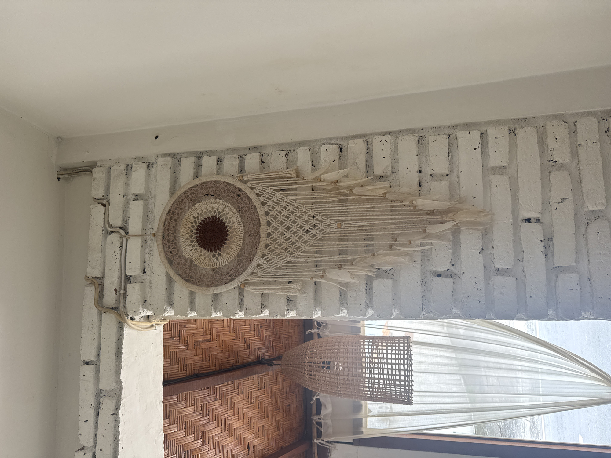 The image shows a portion of the interior of a building in North Kuta, Indonesia. A white-painted brick wall supports a macrame wall hanging featuring a circular woven center and hanging strands. Below, a woven lamp hangs from a ceiling constructed of woven material. Sheer white curtains obscure a window, allowing natural light to permeate the space. An electrical conduit runs along the brick wall.