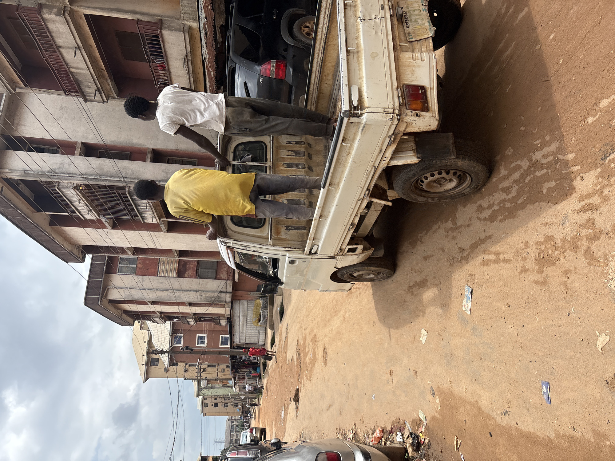 Two young Black males are standing in the bed of a white pickup truck on a dirt road in Onitsha, Nigeria. The male on the left is wearing a yellow t-shirt and grey pants. The male on the right is wearing a white t-shirt and grey pants. Both males are looking forward. The truck is facing to the left, and the bed is empty except for the two men. The road is unpaved and has puddles of water on it. There are buildings on either side of the road, some of which appear to be residential. The sky is overcast.