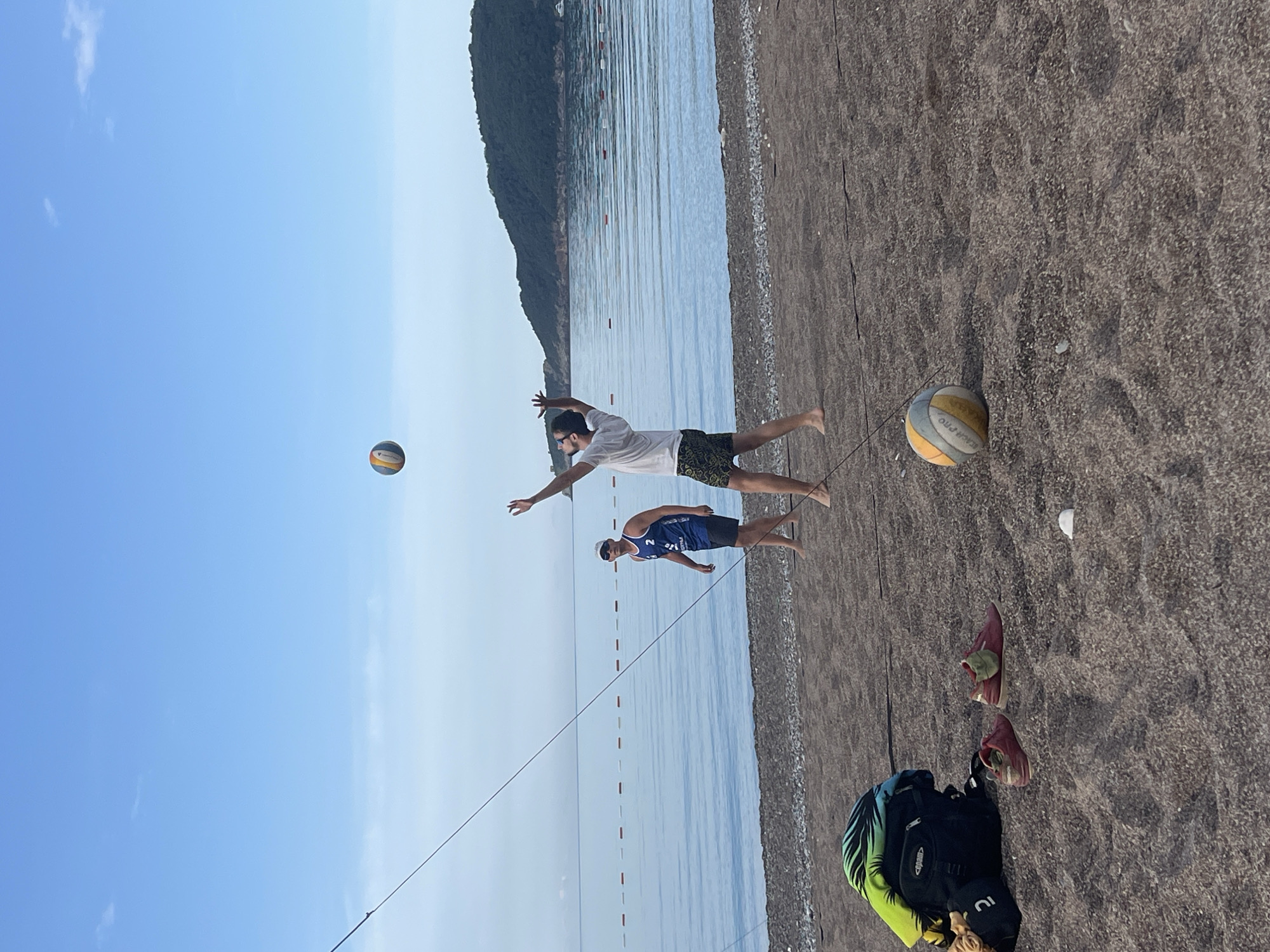 The image depicts a beach volleyball game in Boreti, Montenegro. Two men are playing on a dark sand beach along the Adriatic Sea, with a backdrop of a mountainous coastline under a clear, pale blue sky. One man in a white t-shirt and dark shorts is in mid-air, reaching to hit a volleyball. The second man, wearing a blue jersey and dark shorts, is also positioned on the sand ready to receive the ball. A volleyball lies on the sand near a beach bag and pair of red shoes, suggesting that the players have left their belongings nearby. A volleyball net is set up between the players. Red buoys float in the sea behind them.