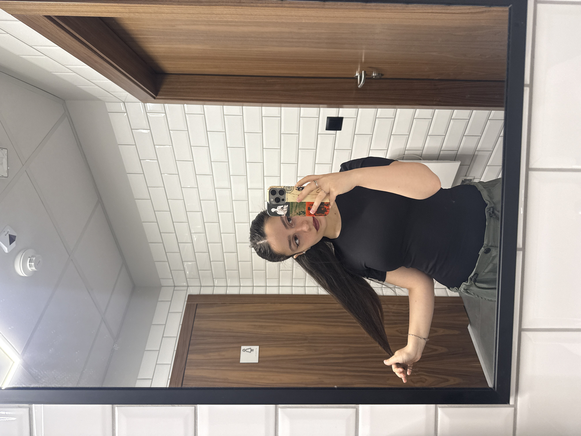 A young woman with dark hair pulled into two pigtails takes a mirror selfie in a public restroom. She is wearing a black t-shirt and green cargo pants. Her left hand is holding her hair while her right hand holds her phone, which has a colorful case. The restroom has white subway tiles on the walls and a wooden door with a women's restroom symbol on it. A smoke detector is visible on the ceiling. The location is identified as Opole, Poland.