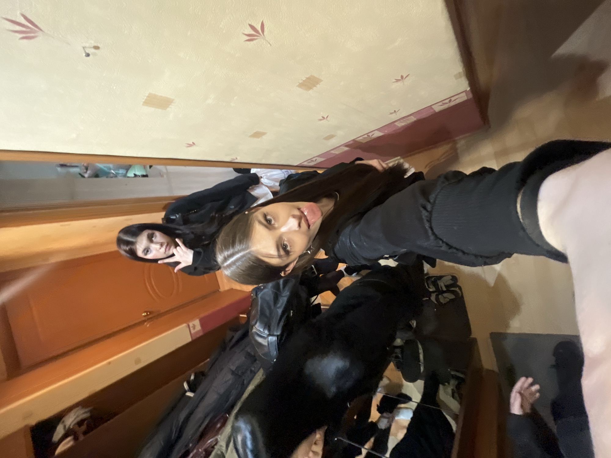 This is a casual, indoor selfie featuring two young women, likely taken in a room or hallway in Vsevolozhsk, Russia. The image is oriented vertically but appears to be taken with the camera held at an upward angle or rotated.
The woman closer to the camera, positioned on the right, has long dark hair and is wearing a dark top or jacket. She looks directly at the camera with a playful expression, her tongue slightly out. Her arm, covered in dark fabric, is visible in the foreground, suggesting she is holding the phone. Behind her, on the left, another young woman with long dark hair and a dark jacket makes a "V" sign with her right hand, also looking towards the camera.
The background features a light-colored (beige/cream) wall adorned with a repeating pattern of small, reddish-brown leaf-like motifs. Several rectangular patches of what appears to be tape or adhesive are visible on the wall. A wooden trim runs along the lower portion of the wall. To the left, a wooden door with a distinctive circular panel design and a doorknob is partially visible. Below the door, a section of wooden furniture, possibly an open wardrobe or shelving unit, contains dark clothing items. A reflective surface, likely a mirror, also captures faint reflections.
The activity is a casual photo session, possibly a selfie among friends. The expressions and gestures of the subjects convey a sense of playfulness and camaraderie. The lighting suggests artificial indoor illumination. No discernible text is visible within the image.