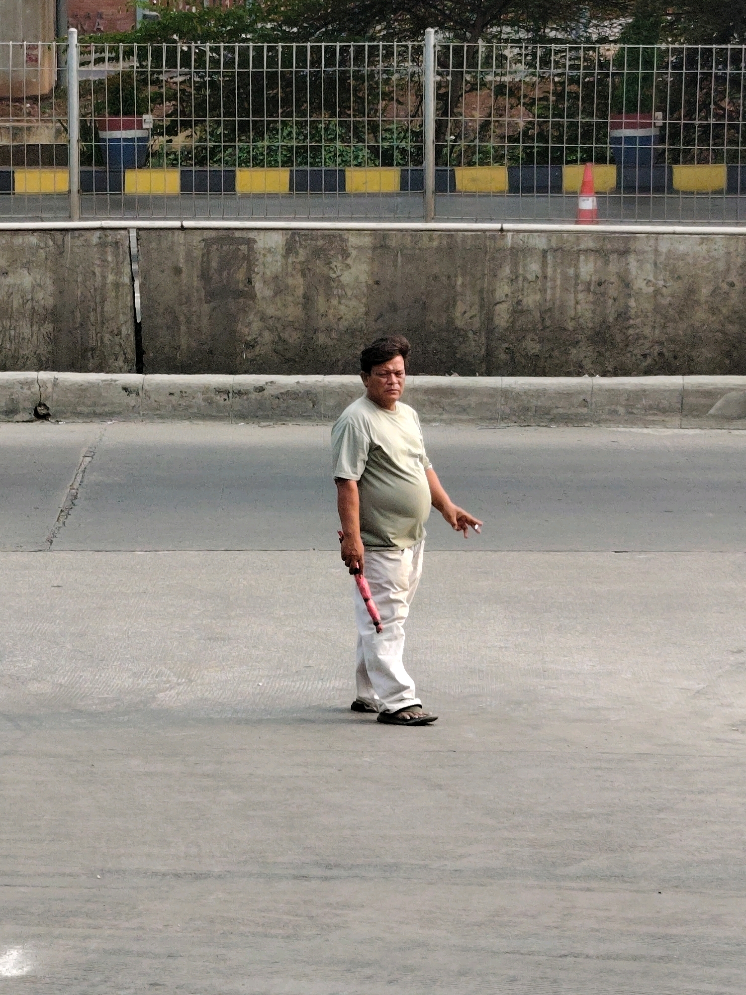 A medium-shot photograph shows a man standing in an outdoor urban environment, likely on a road or pavement. The man, who appears to be of South Asian descent, is wearing a light olive green t-shirt and loose-fitting white pants. He is holding a red object in his right hand and appears to be smoking a cigarette with his left hand. His stance suggests he is looking off to his left.
The background features a grey concrete wall, and behind that, a metal fence with a yellow and black striped barrier. Beyond the fence, trees and foliage are visible. A bright orange traffic cone with white reflective bands is positioned in the background to the right of the frame. The ground the man is standing on is a light grey paved surface with some textured lines.
The lighting suggests it is daytime, with a harsh, bright sun casting shadows. The overall scene conveys a casual, potentially everyday moment captured in an urban setting, possibly near a road or a construction area, given the traffic cone and the utilitarian nature of the background elements. The location is identified as Bekasi, Indonesia.