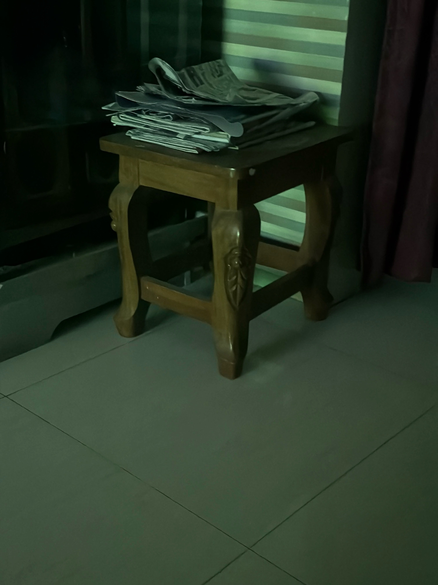 In Bandar, Bangladesh, a low-angle shot captures a small, ornate wooden stool with stacked papers on top. The stool has four curved legs and decorative carvings on its sides. The papers, appearing to be newspapers or magazines, are fanned out and loosely piled. Behind the stool, a wall with horizontal stripes in muted green, blue, and beige tones is visible, along with a dark purple curtain on the right. The floor is tiled with large, light gray tiles. The lighting is dim, casting a green hue over the scene, and suggesting an indoor environment.