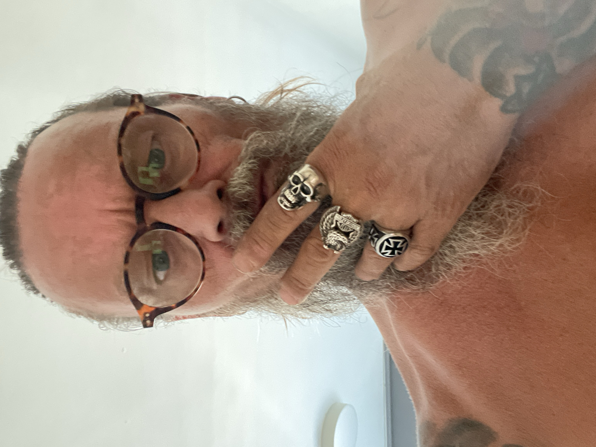This close-up, horizontally oriented media file features a person with a beard and eyeglasses. The person is wearing multiple rings on their fingers, including a skull ring and a ring with a cross design. A tattoo is visible on their arm. The background is a plain, light-colored wall, suggesting an indoor setting. The lighting appears to be natural, possibly from a window, and there are no obvious indications of the time of day or weather conditions. The person's face is partially obscured by their hand and beard, and their expression is not clearly discernible. There is no visible text in the image. The media file was taken in Netanya, Israel.