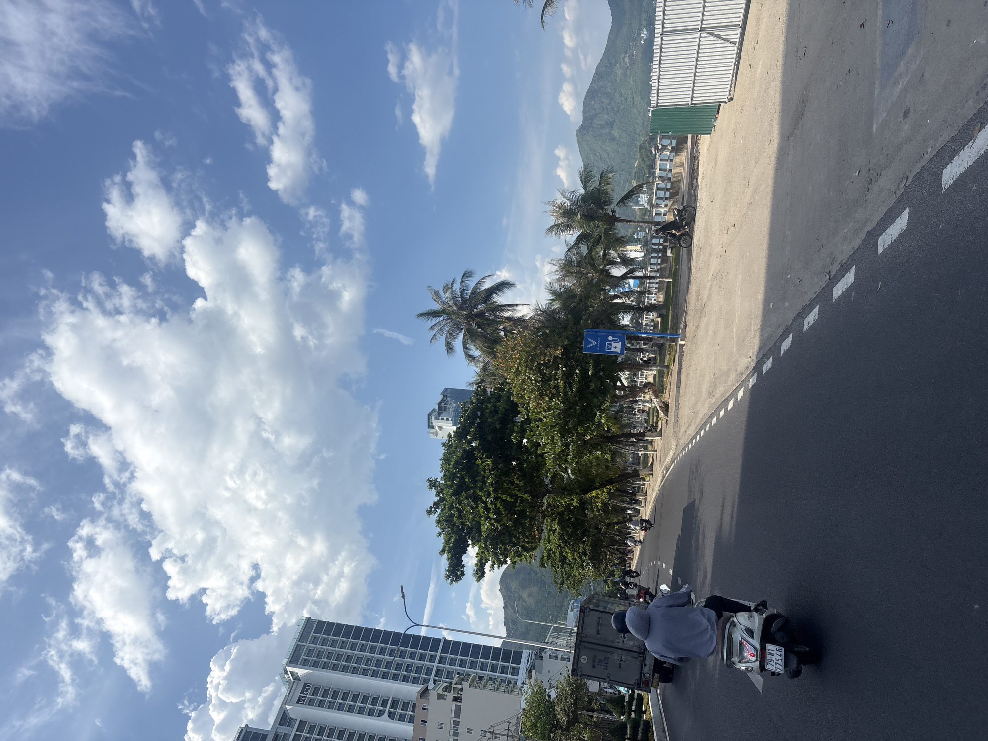 The image shows a scene in Nha Trang, Vietnam, likely along a coastal road. In the foreground, a person is riding a white scooter on the right side of a paved road. The rider is wearing a grey hooded sweatshirt. Behind the scooter, a grey truck is visible on the left side of the road. Further down the road, several other people on motorcycles are also traveling.
The environment includes tall, modern buildings on the left side of the road, appearing to be high-rise apartments or hotels. On the right side of the road, there is a sandy area with palm trees and other greenery, leading towards a metal fence. In the background, a lush green mountain range is visible under a bright blue sky with scattered white clouds. The road has dashed white lines marking its lanes. The overall impression is a sunny day in a developed, coastal city.