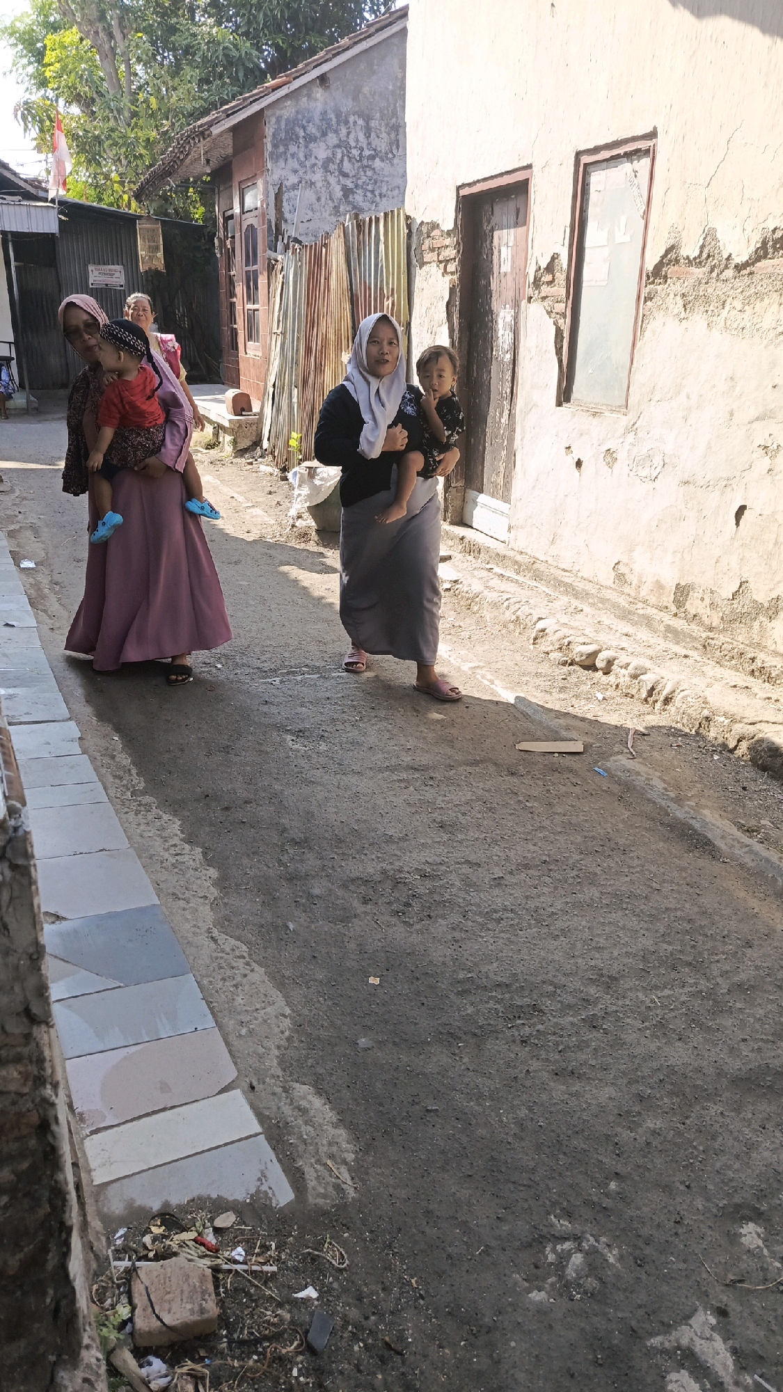 In the city of Pekauman Kulon, Indonesia, a narrow, unpaved alleyway is captured under bright sunlight. Two women, both wearing hijabs, are walking towards the viewer. The woman on the left carries a young child in her arms, dressed in a red shirt and dark pants, with blue sandals. She wears a patterned top and a long, mauve skirt. The woman on the right, in a black top and a long grey skirt, also holds a young child, who is wearing a dark patterned garment.
The alley is lined with buildings. The building on the right has a weathered, light-colored exterior with a wooden door and a window with obscured glass, showing signs of deterioration. The building on the left is partially visible, with corrugated metal siding and what appears to be a section of an Indonesian flag hanging. The ground of the alley is rough and uneven, with scattered debris, including a piece of cardboard and some plastic. A stone walkway or curb runs along the left side of the alley. Trees with green foliage are visible in the background, suggesting a warm climate. The overall impression is one of everyday life in a densely populated urban environment.