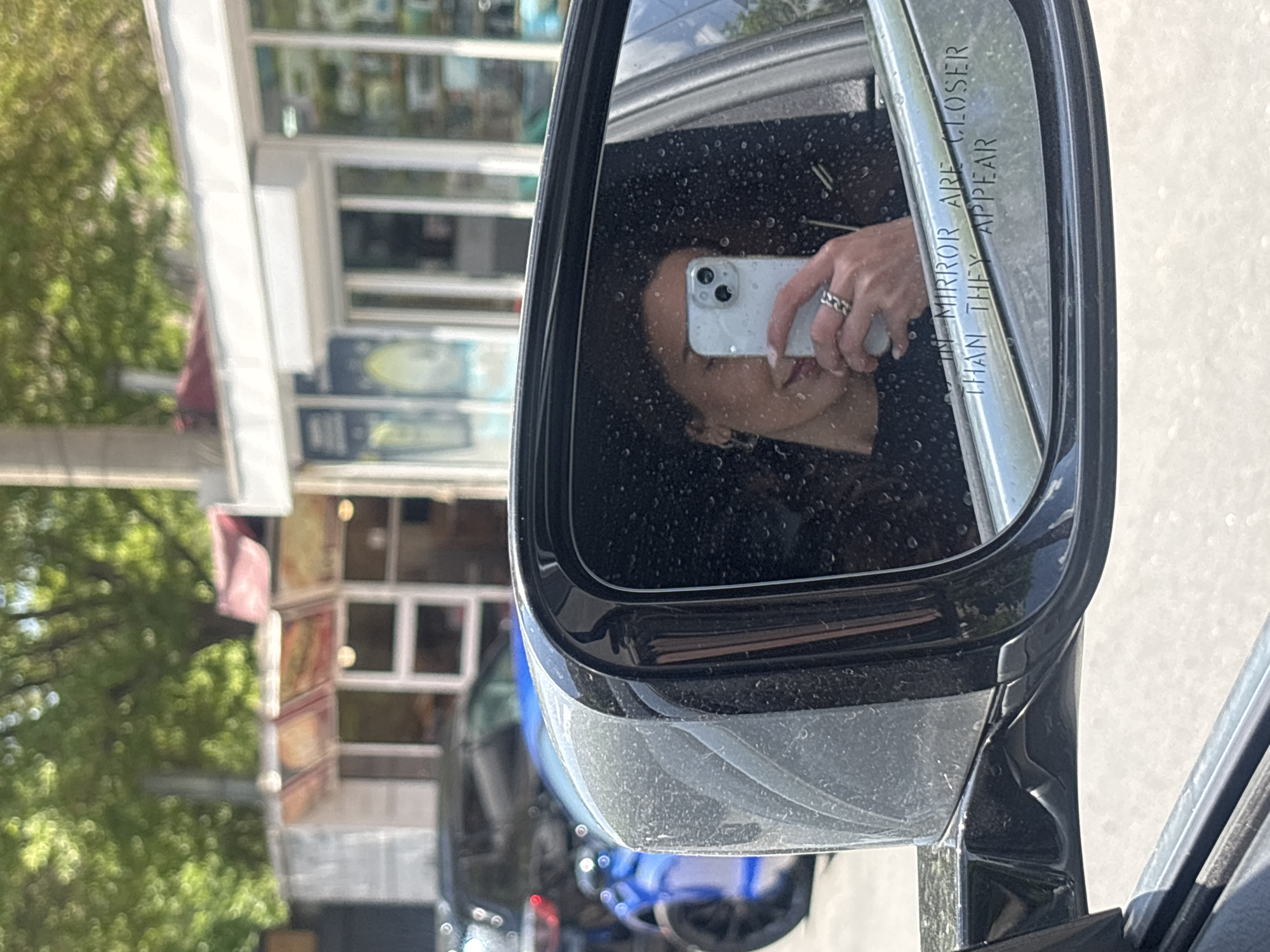 The image captures a person reflected in a car's side mirror. The person is holding a smartphone and appears to be taking a picture of themselves in the mirror. The mirror itself bears the standard safety warning, "Objects in mirror are closer than they appear." The surroundings reflected in the mirror and visible in the background suggest an urban environment, with buildings, trees, and other vehicles present. However, it is not possible to determine that the location is in the city of Yerevan, Armenia.