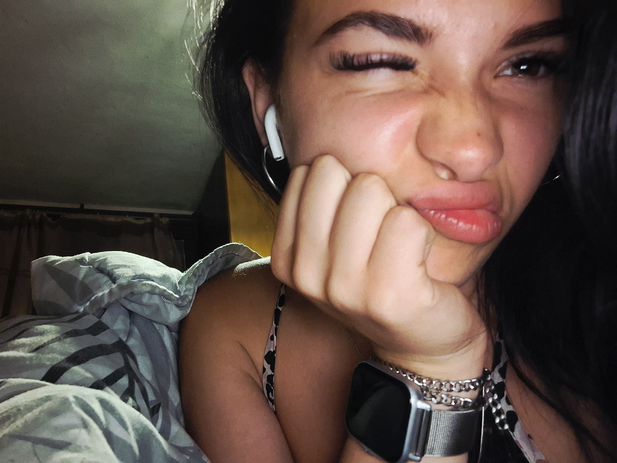 A close-up, dimly lit image shows a young woman playfully winking at the camera while lying down, possibly in bed. She has dark hair and is seen from the chest up. Her right eye is wide open, looking towards the viewer, while her left eye is tightly winked. Her nose is scrunched, and her lips are pursed in a puckering expression, with her tongue slightly visible. Her right hand is propping up her chin.
She is wearing a white earbud in her left ear, a silver hoop earring, and a smartwatch on her left wrist, accompanied by multiple chain bracelets. A patterned top, possibly an animal print, is visible on her shoulder and chest. She is partially covered by a light-colored duvet or blanket with a dark striped pattern. The background shows a dark ceiling with some light reflections and dark curtains in the distance, suggesting an indoor setting, likely a bedroom, during the evening or night. No text is visible in the image. The location context provided is Krapets, Bulgaria.